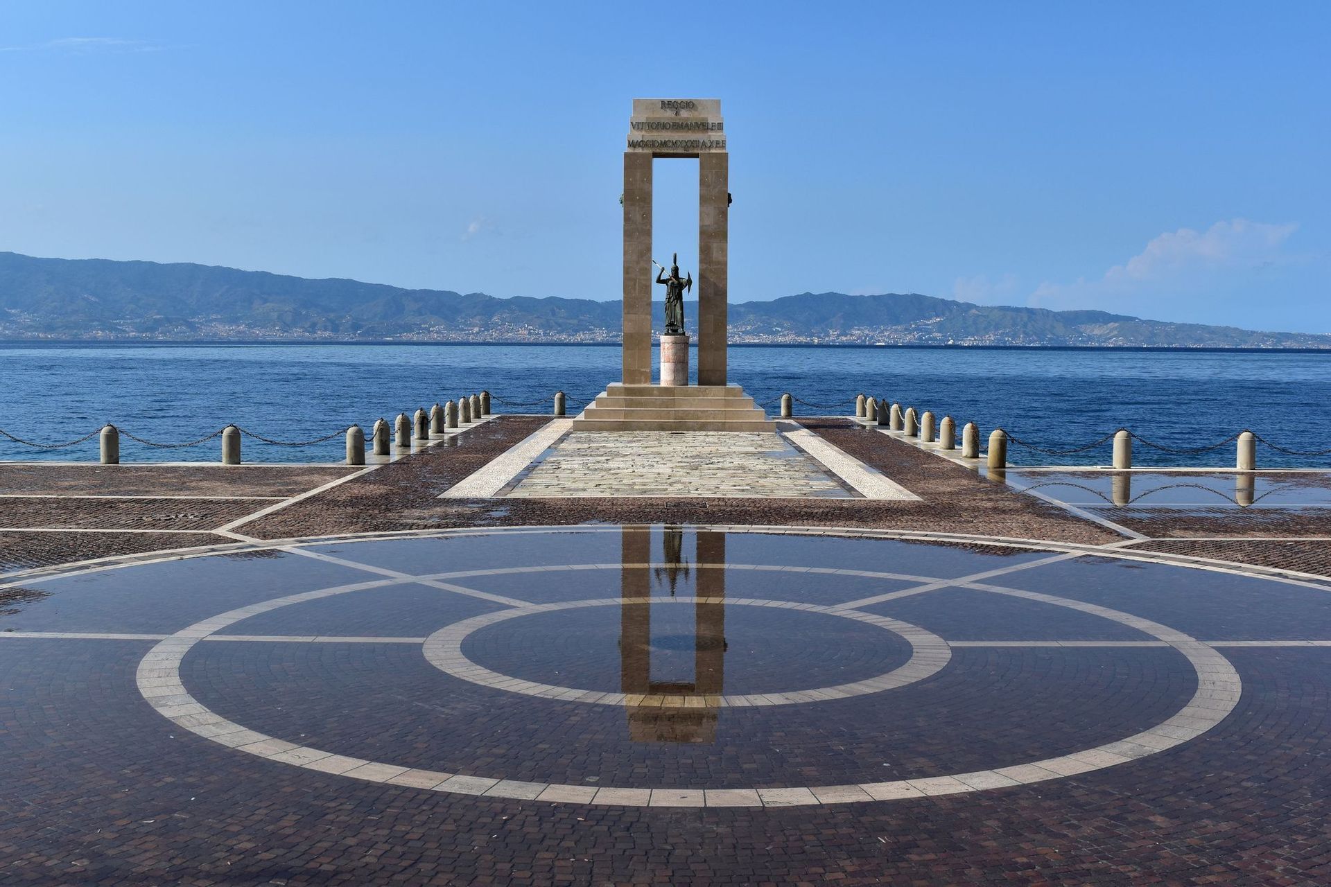 Un monumento su un molo di pietra si riflette in una grande pozzanghera, con il mare e le montagne sullo sfondo.