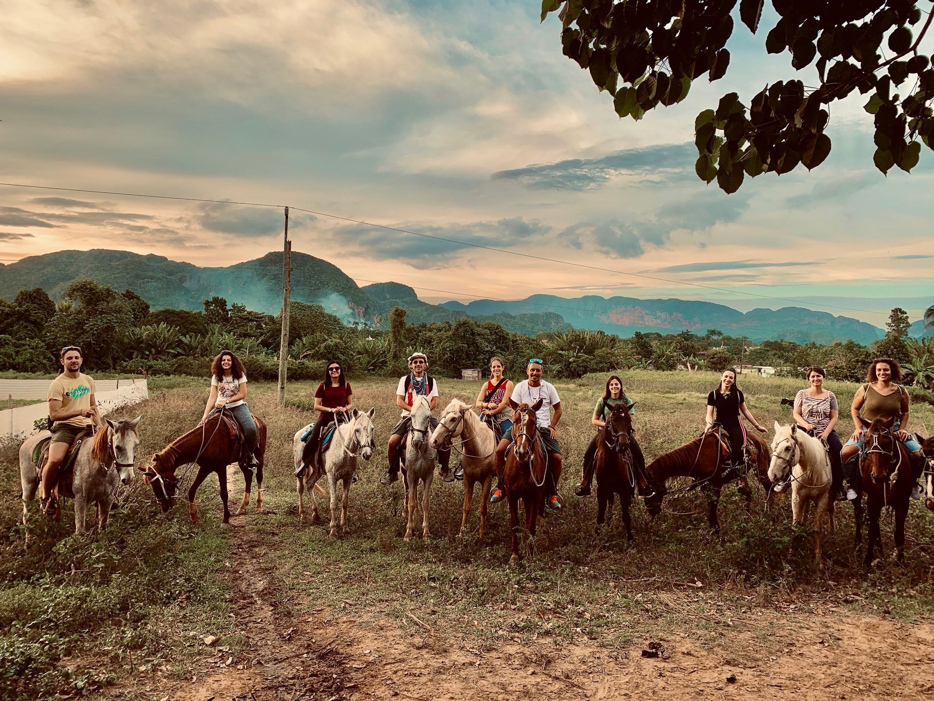 Un groupe WeRoad en voyage pose à cheval dans un champ herbeux avec des montagnes vertes en arrière-plan sous un ciel nuageux.