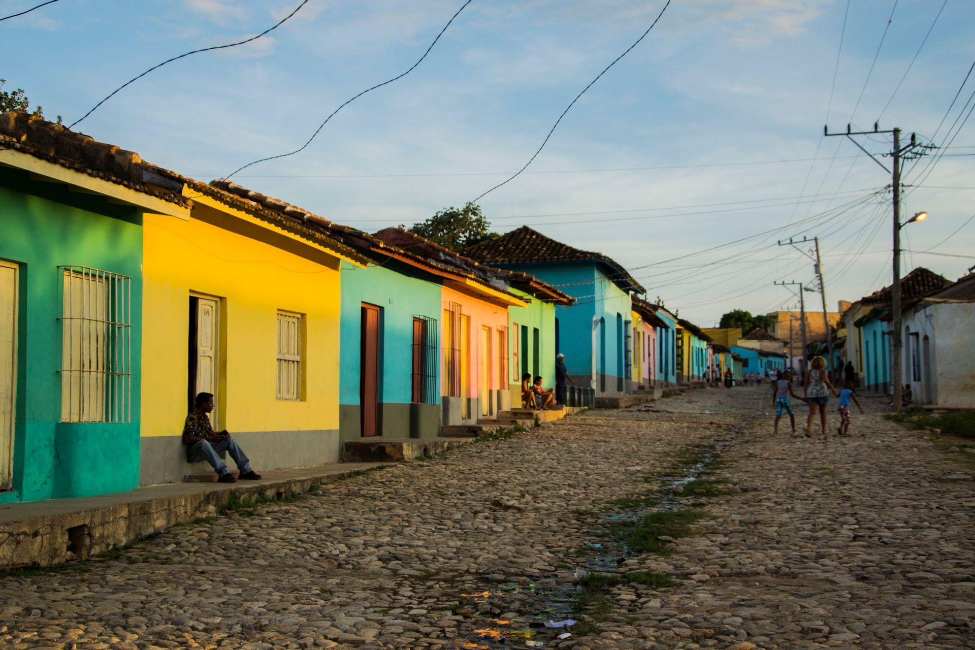 Una calle empedrada bordeada de casas coloridas en verde, amarillo y azul, con gente relajándose al aire libre durante el atardecer.