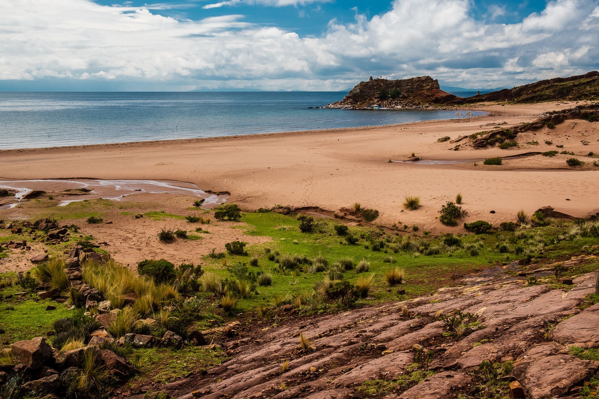 Una playa amplia y arenosa junto a un cuerpo de agua tranquilo, con zonas de césped verde y un promontorio rocoso bajo un cielo parcialmente nublado.
