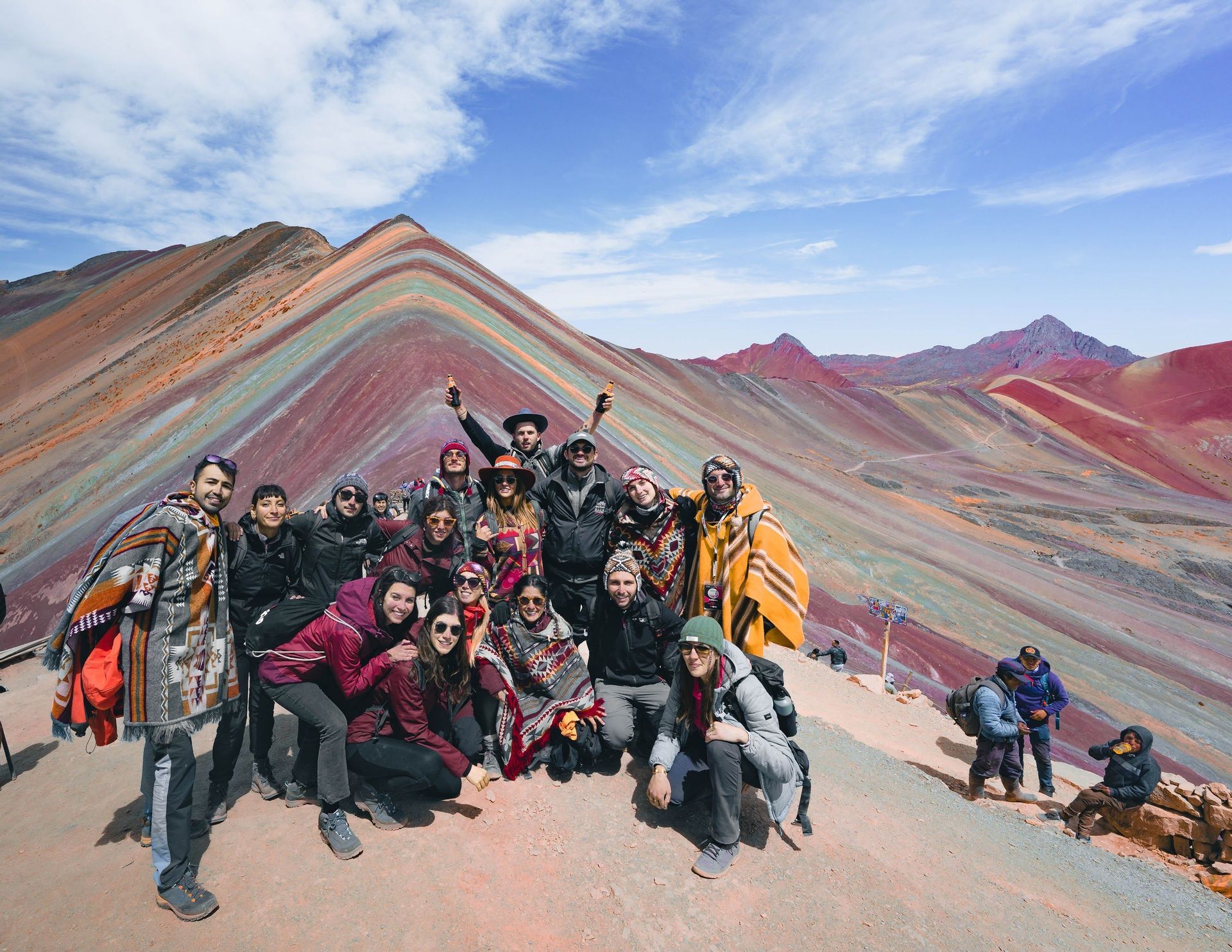 A WeRoad group trip poses for a photo on a mountainside with a large, colorful, striped mountain in the background under a blue sky.