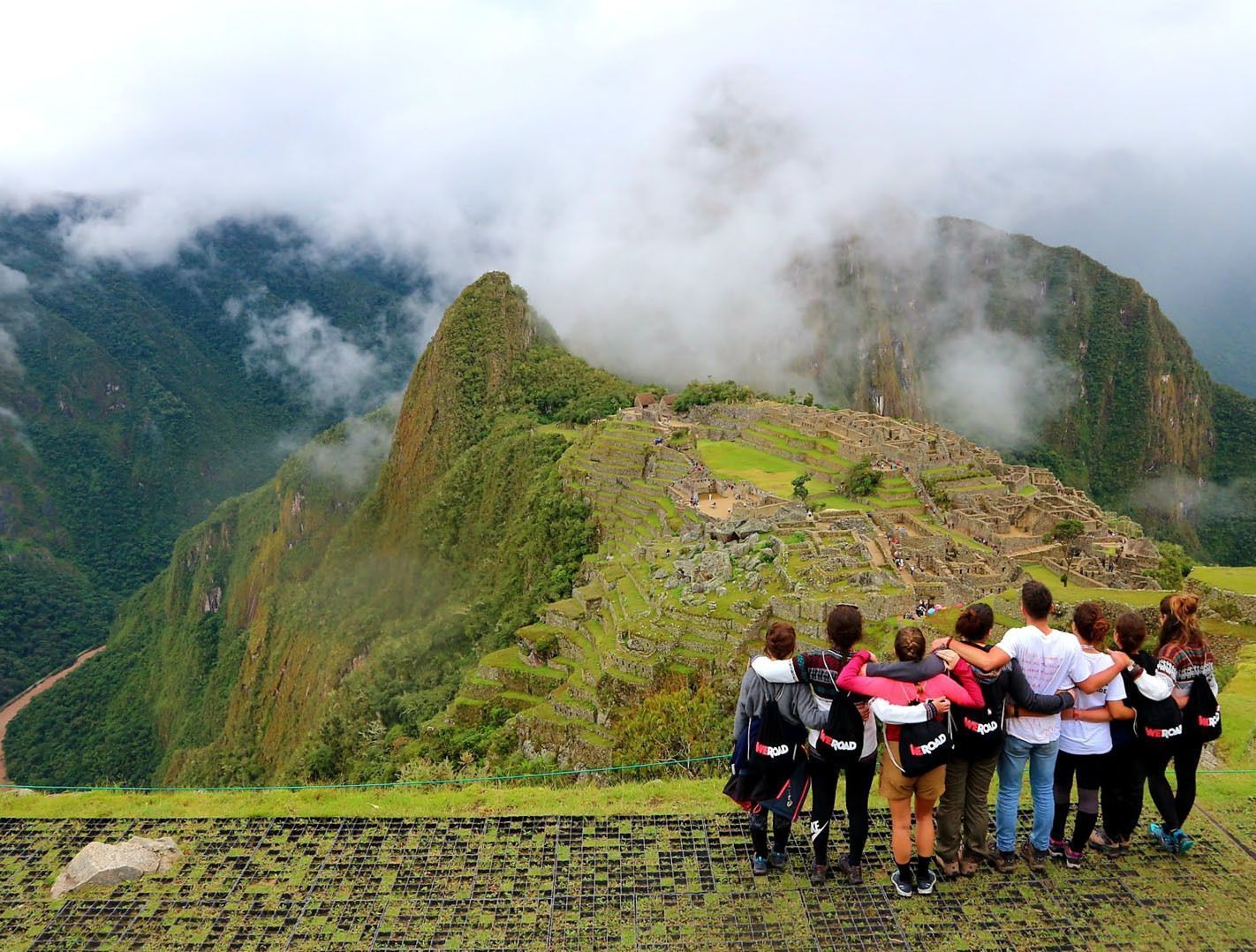 Un grupo de viaje de WeRoad, abrazados, contempla ruinas antiguas en una ladera verde y brumosa desde un mirador.