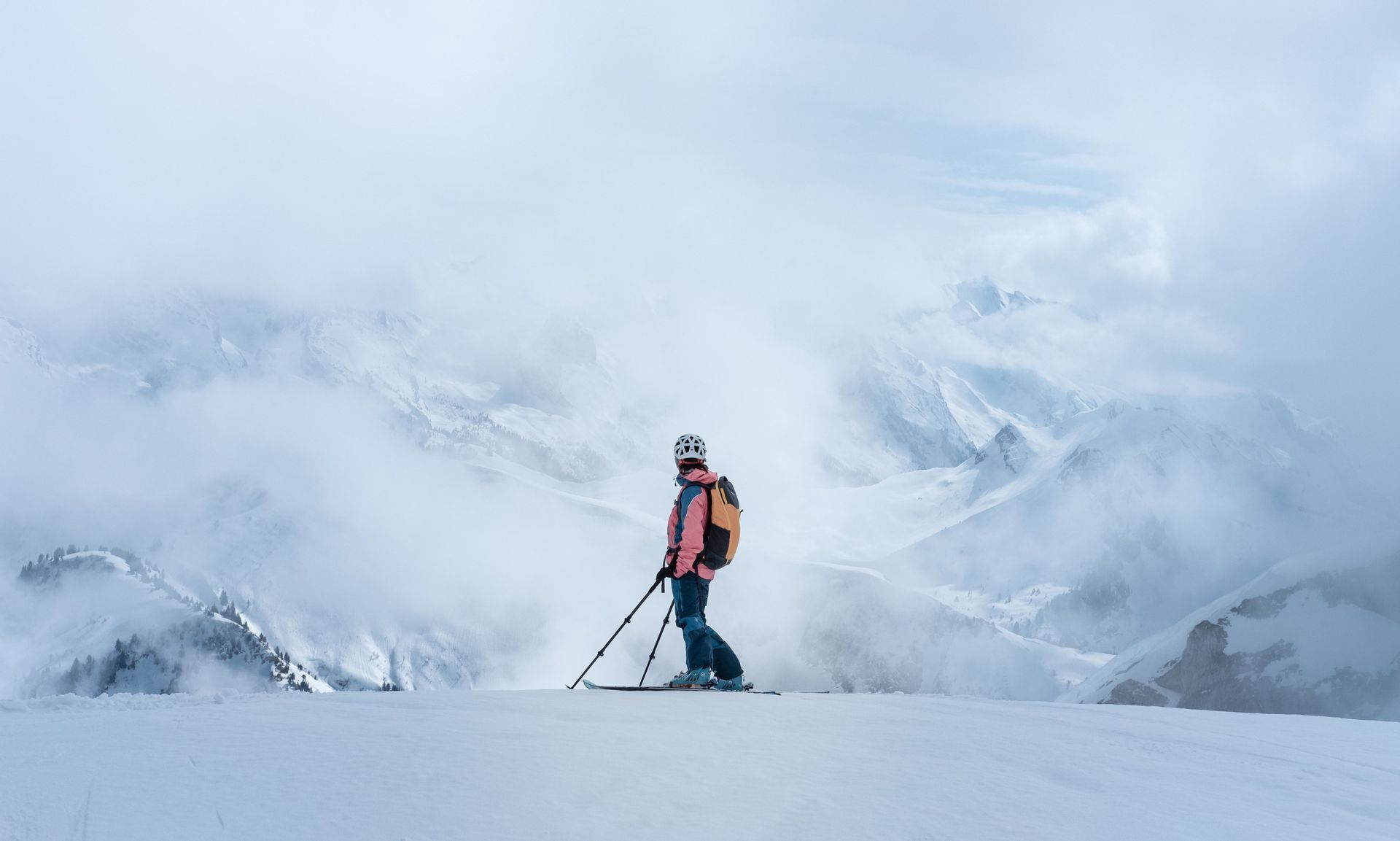 Una persona con gli sci, casco e zaino, si trova su un crinale innevato, ammirando una vasta catena montuosa avvolta dalle nuvole.