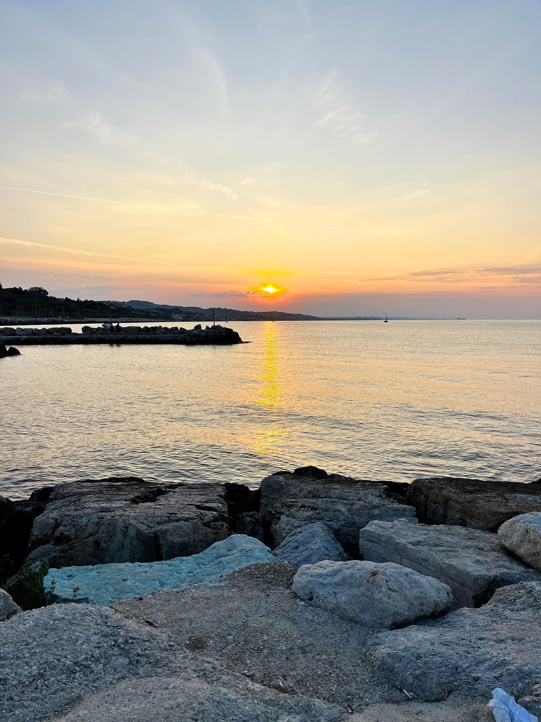 Il sole tramonta su una distesa d'acqua calma, osservato da un frangiflutti roccioso, proiettando un riflesso dorato sulla superficie.