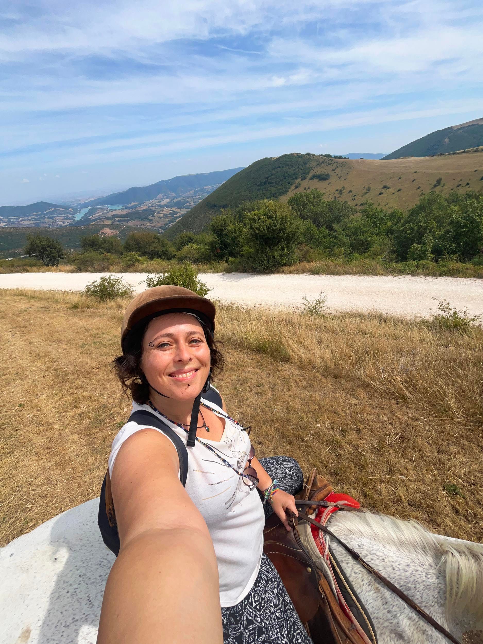 Una donna sorridente con un casco si scatta un selfie mentre cavalca un cavallo bianco su una collina, con una valle e un lago sullo sfondo.