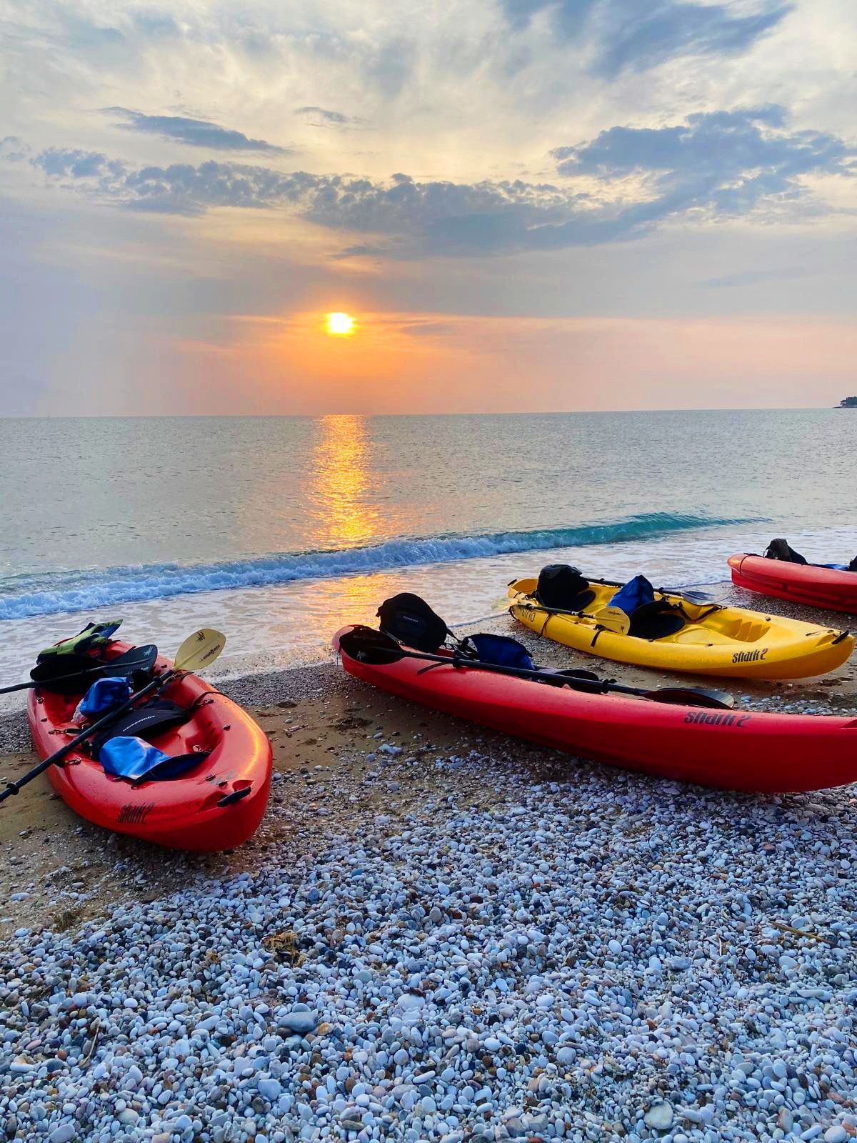 Kayak rossi e gialli riposano su una spiaggia di ciottoli con onde dolci mentre il sole tramonta sull'oceano sotto un cielo nuvoloso.