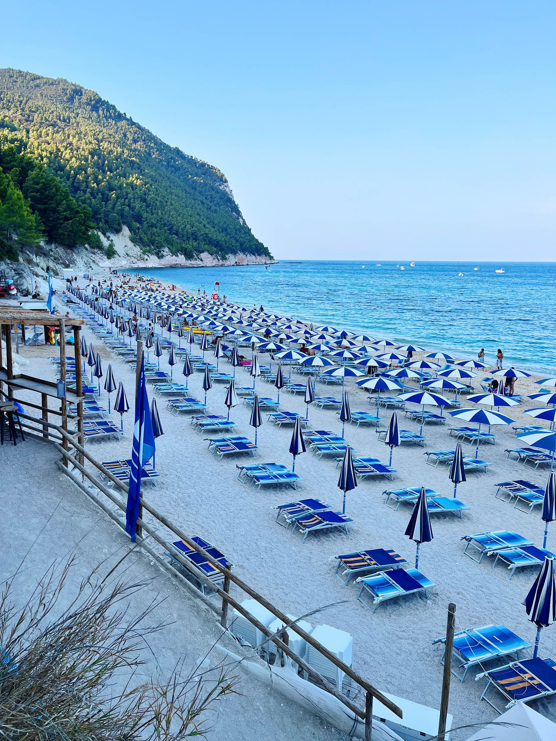 File di ombrelloni e lettini da spiaggia blu e bianchi costeggiano una spiaggia sabbiosa vicino al mare, con una collina boscosa sullo sfondo.