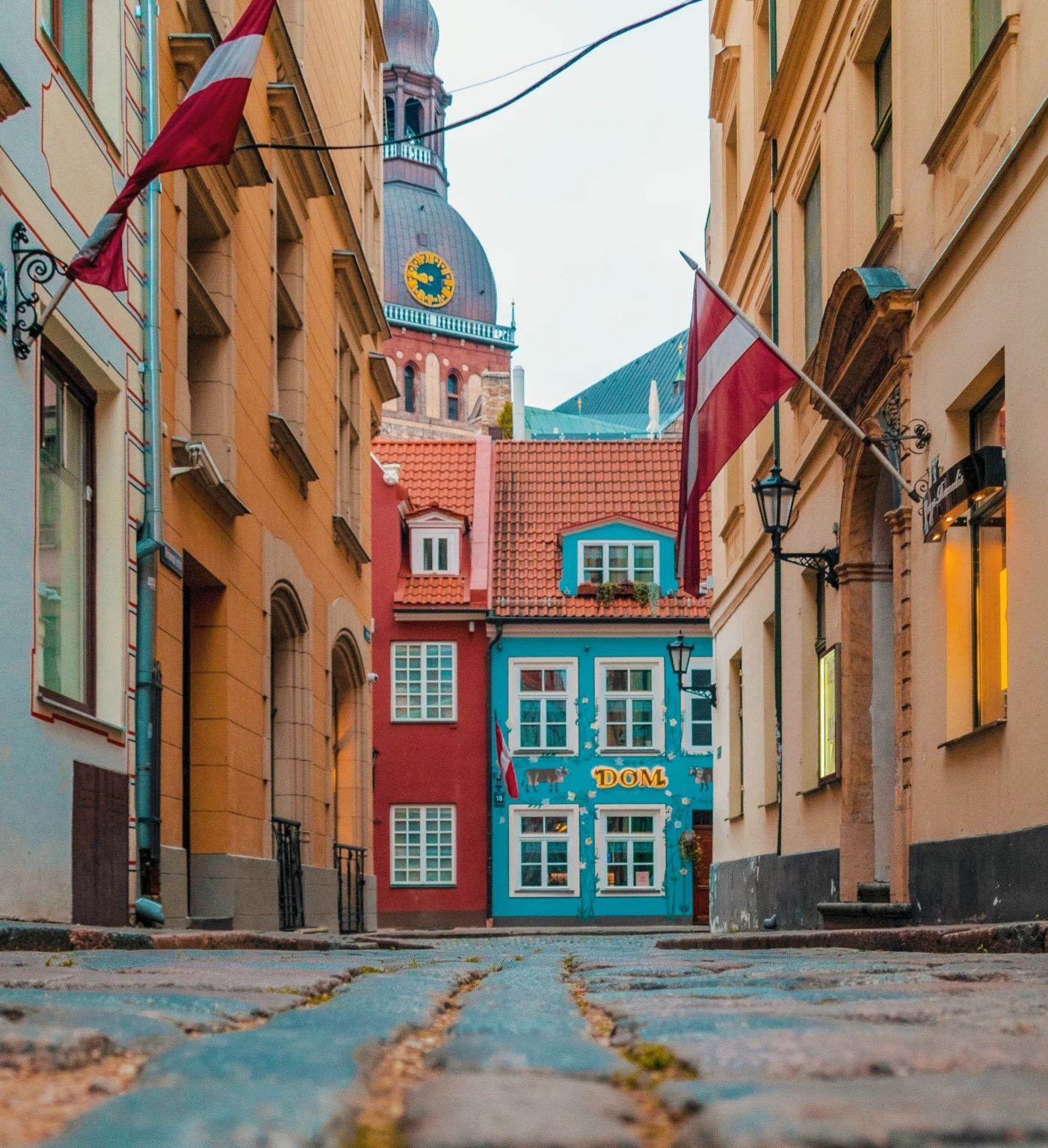Una vista dal basso di una stretta strada acciottolata che porta a edifici colorati, con due bandiere lettoni appese e una torre dell'orologio sullo sfondo.