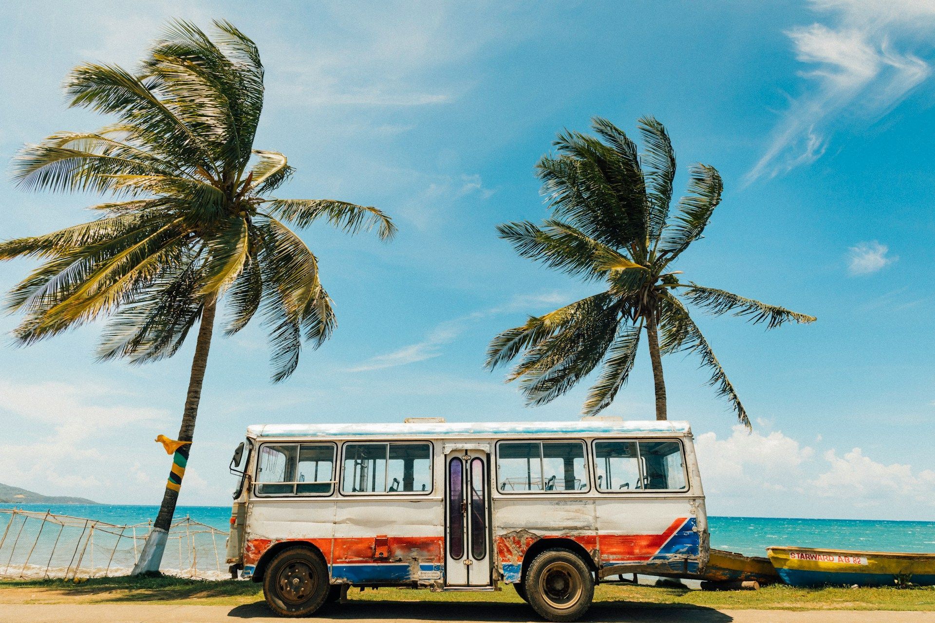Un vecchio autobus parcheggiato su una strada accanto a una spiaggia tropicale, con due grandi palme che ondeggiano contro un cielo blu.