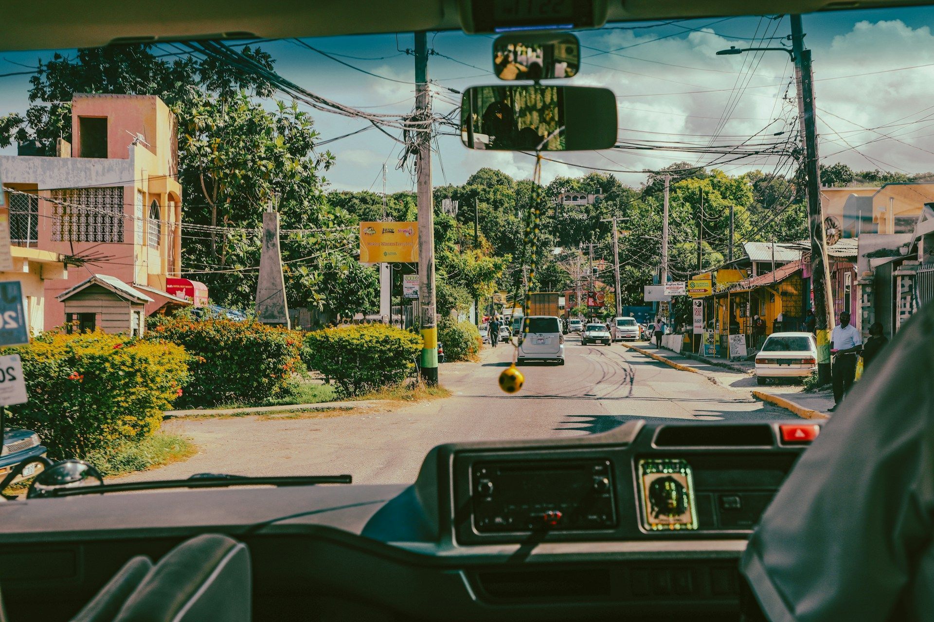 Vista dal cruscotto di un'auto su una strada di città con auto, edifici colorati e alberi verdi rigogliosi.