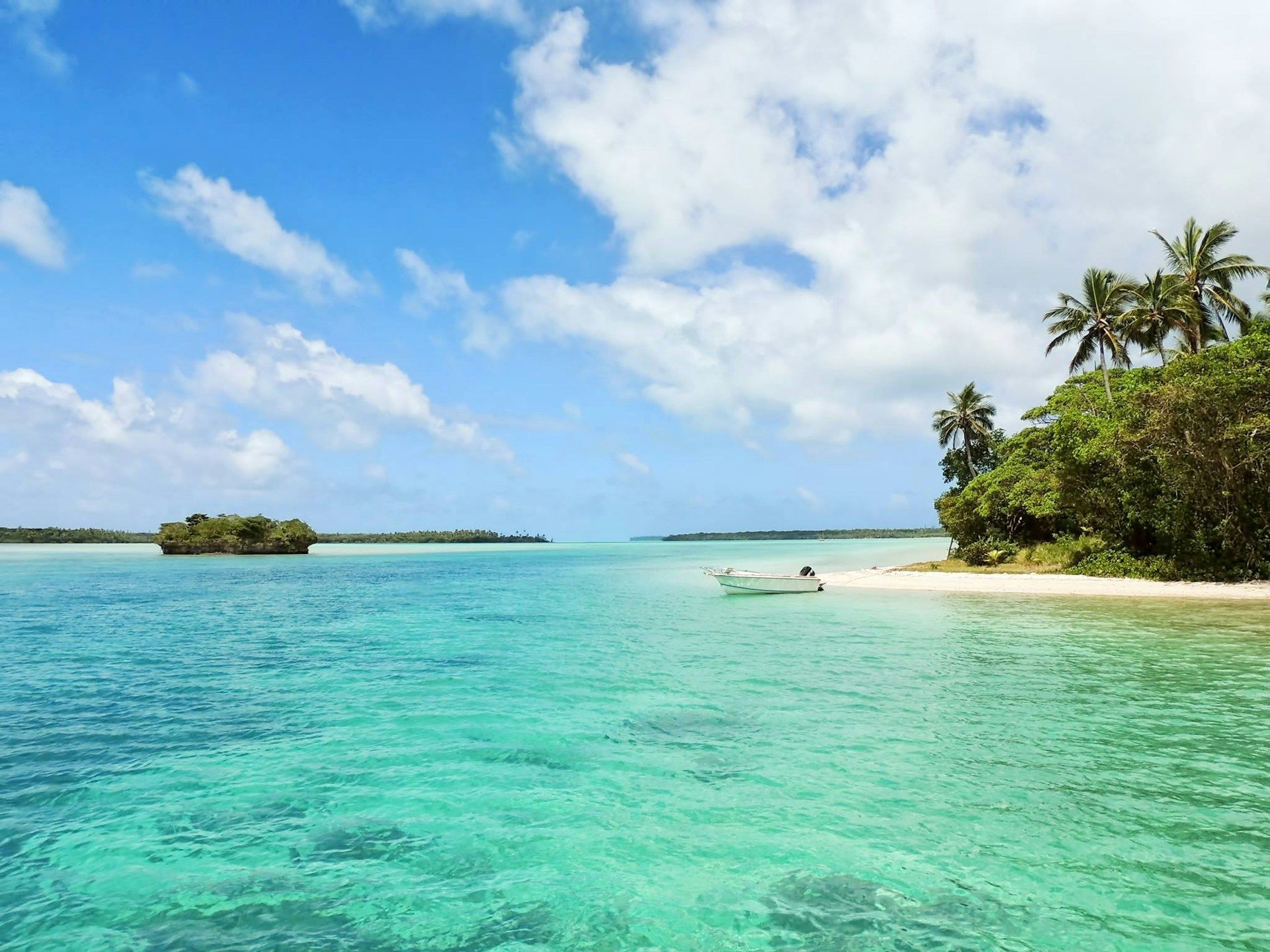 Una piccola barca è ormeggiata sulla spiaggia sabbiosa di un'isola tropicale con acqua cristallina e turchese sotto un cielo parzialmente nuvoloso.