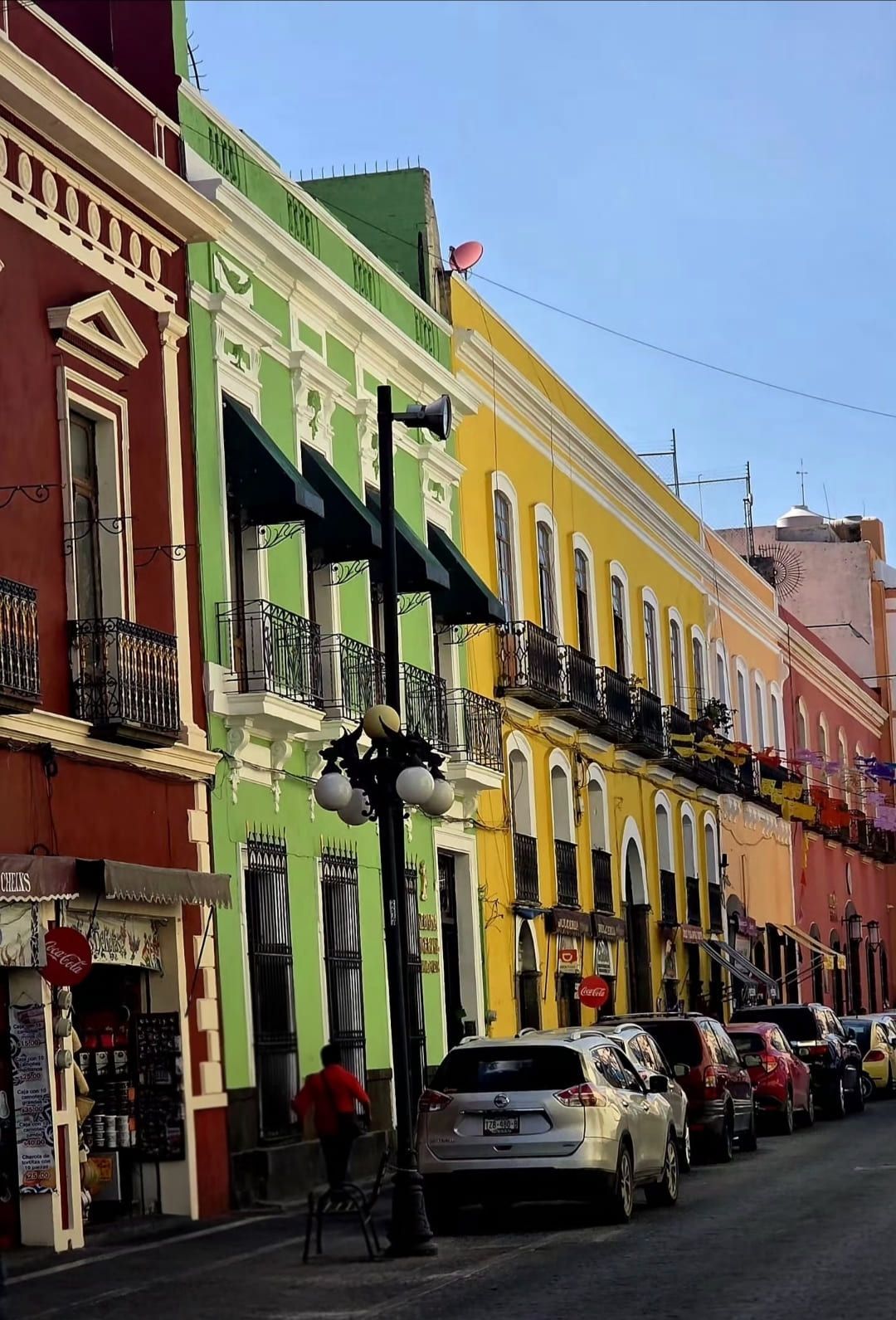 Una fila de coloridos edificios rojos, verdes y amarillos con balcones bordea una calle estrecha con coches estacionados.
