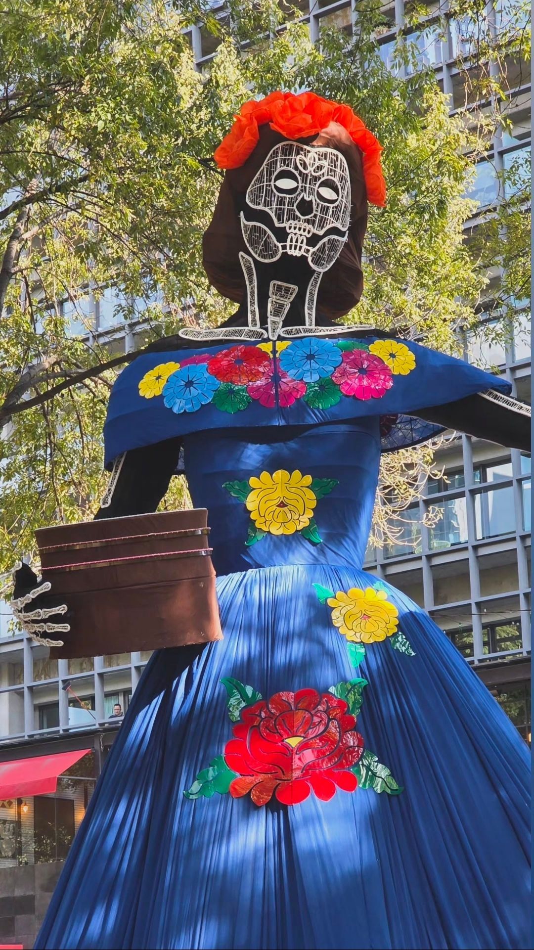 Una gran figura de Calavera Catrina con un rostro de calavera blanco y un vestido azul adornado con flores coloridas se encuentra al aire libre.