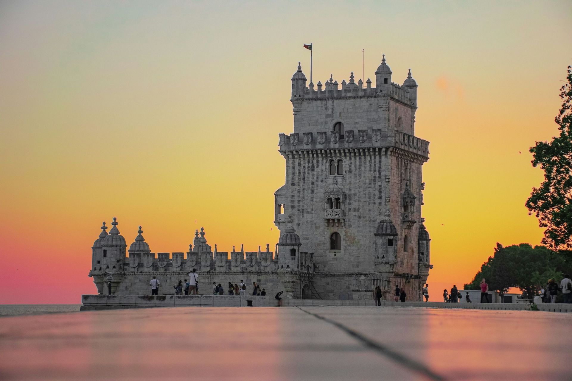 Una fortezza con torre in pietra ornata si erge sul lungomare al tramonto, vista da un'angolazione bassa su una piazza pavimentata.
