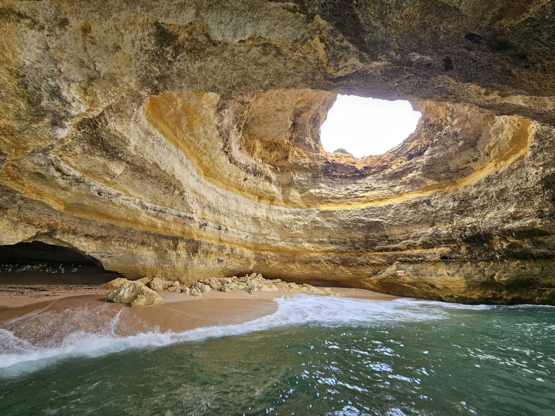 Le onde accarezzano una piccola spiaggia sabbiosa all'interno di una grande grotta marina, dotata di un'apertura rotonda verso il cielo nel suo soffitto.