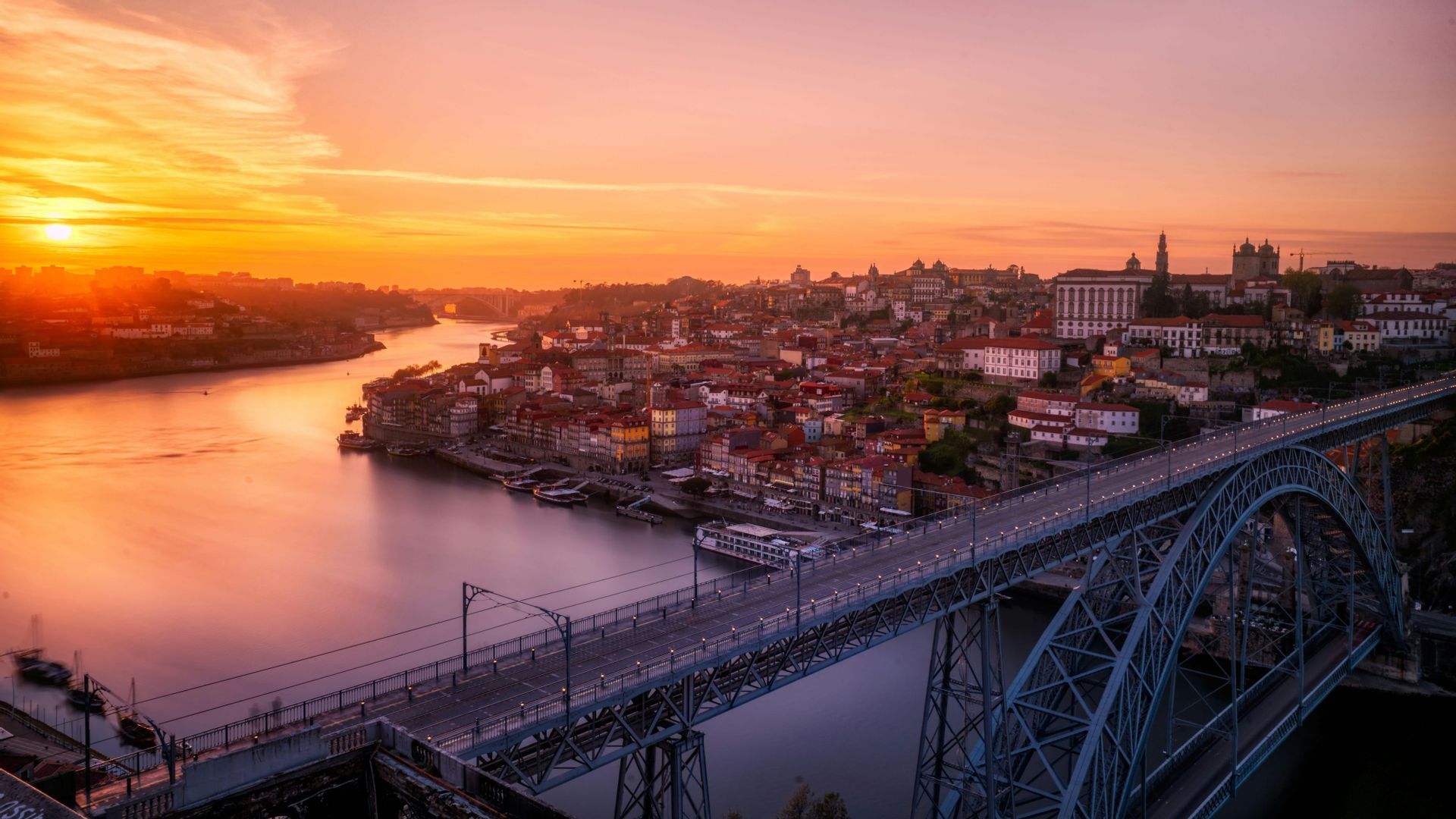 Una veduta aerea di un grande ponte ad arco su un fiume, accanto a una città, durante un tramonto arancione vibrante.