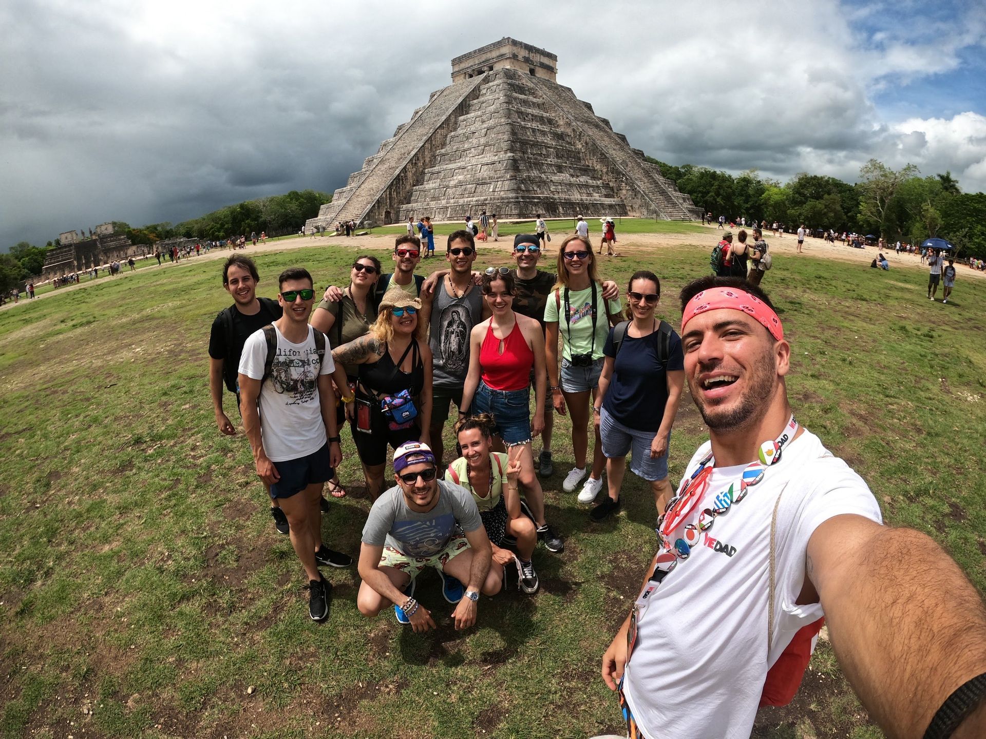 A WeRoad group trip takes a smiling selfie on a grassy field in front of a large ancient stone pyramid under a cloudy sky.