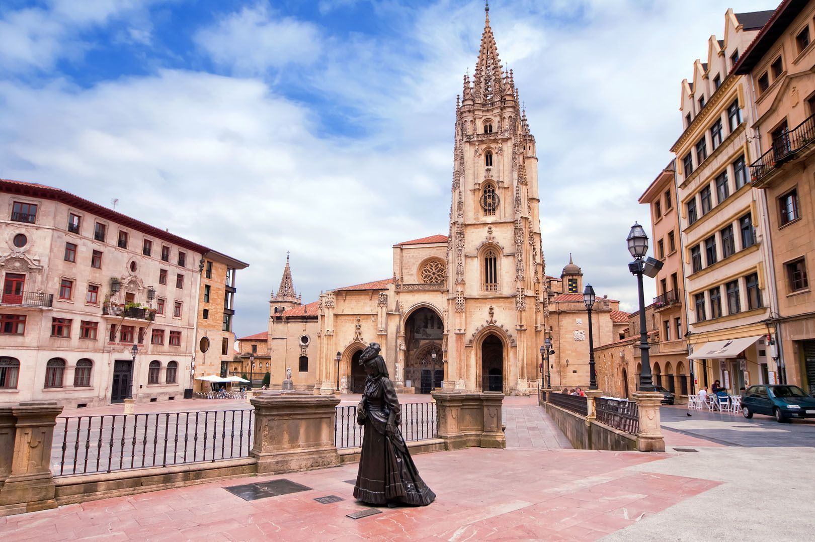Una estatua de bronce de una mujer se alza en una plaza pavimentada frente a una ornamentada catedral gótica bajo un cielo parcialmente nublado.