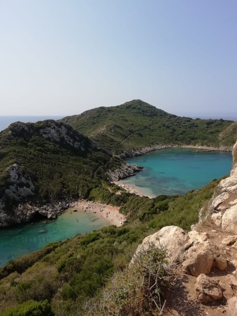 Vue plongeante sur une double crique aux eaux turquoise et plages de sable, nichée entre des collines verdoyantes sous un ciel clair.