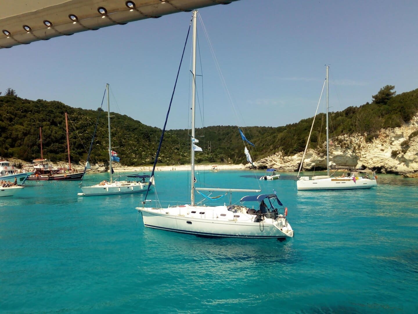 Sailboats anchored in a calm bay with turquoise water, a sandy beach, and green, tree-covered hills.