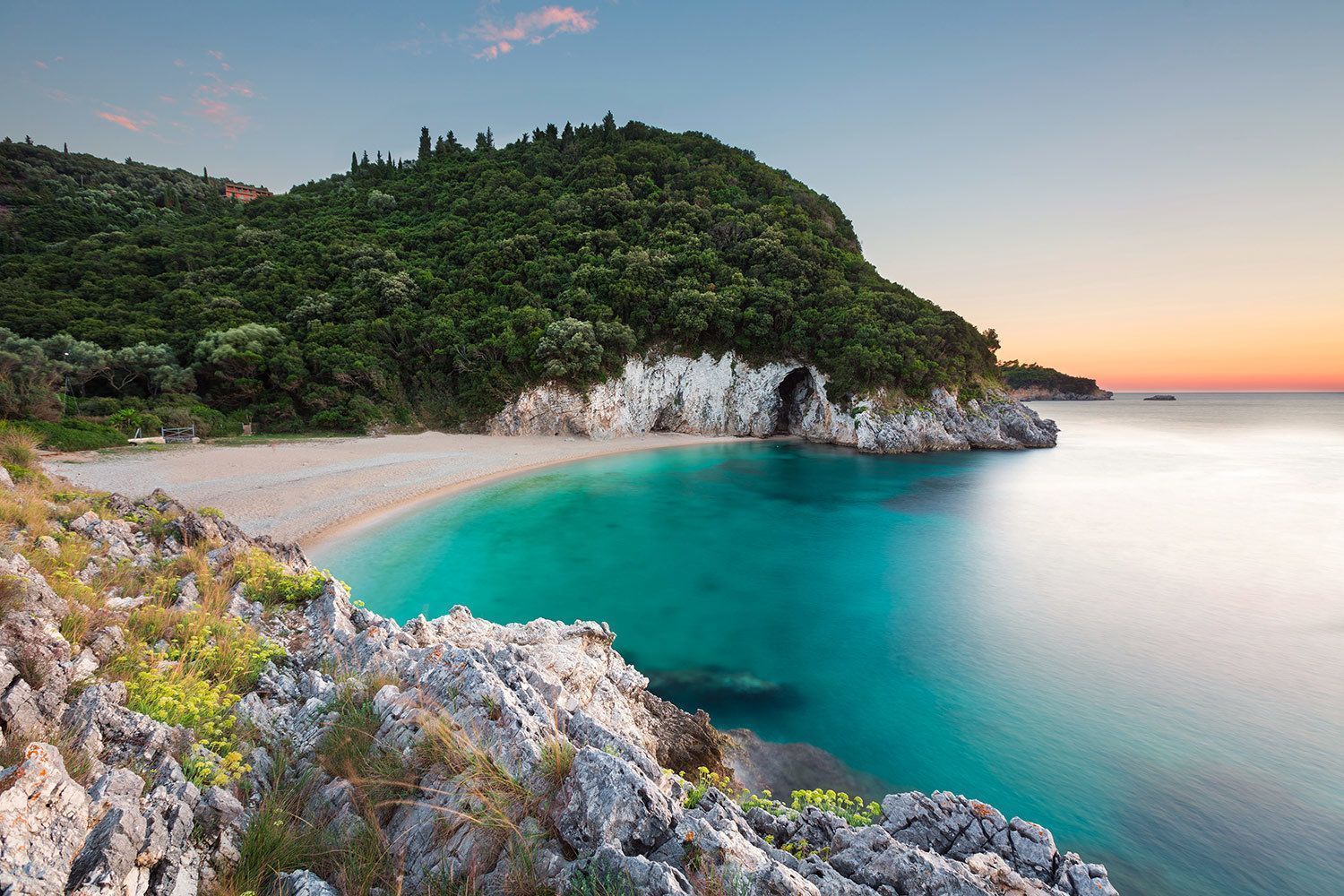 A secluded beach with turquoise water sits in a cove with a sea cave, nestled below a forested hill at sunset.