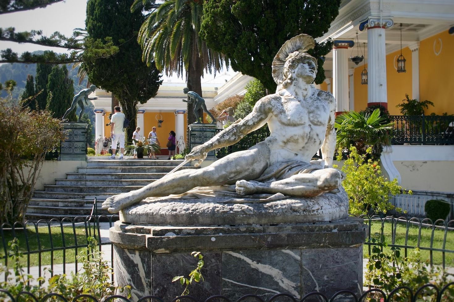 Una statua di marmo logorata raffigurante un guerriero reclinato in un giardino lussureggiante, con un palazzo giallo e colonne bianche sullo sfondo.