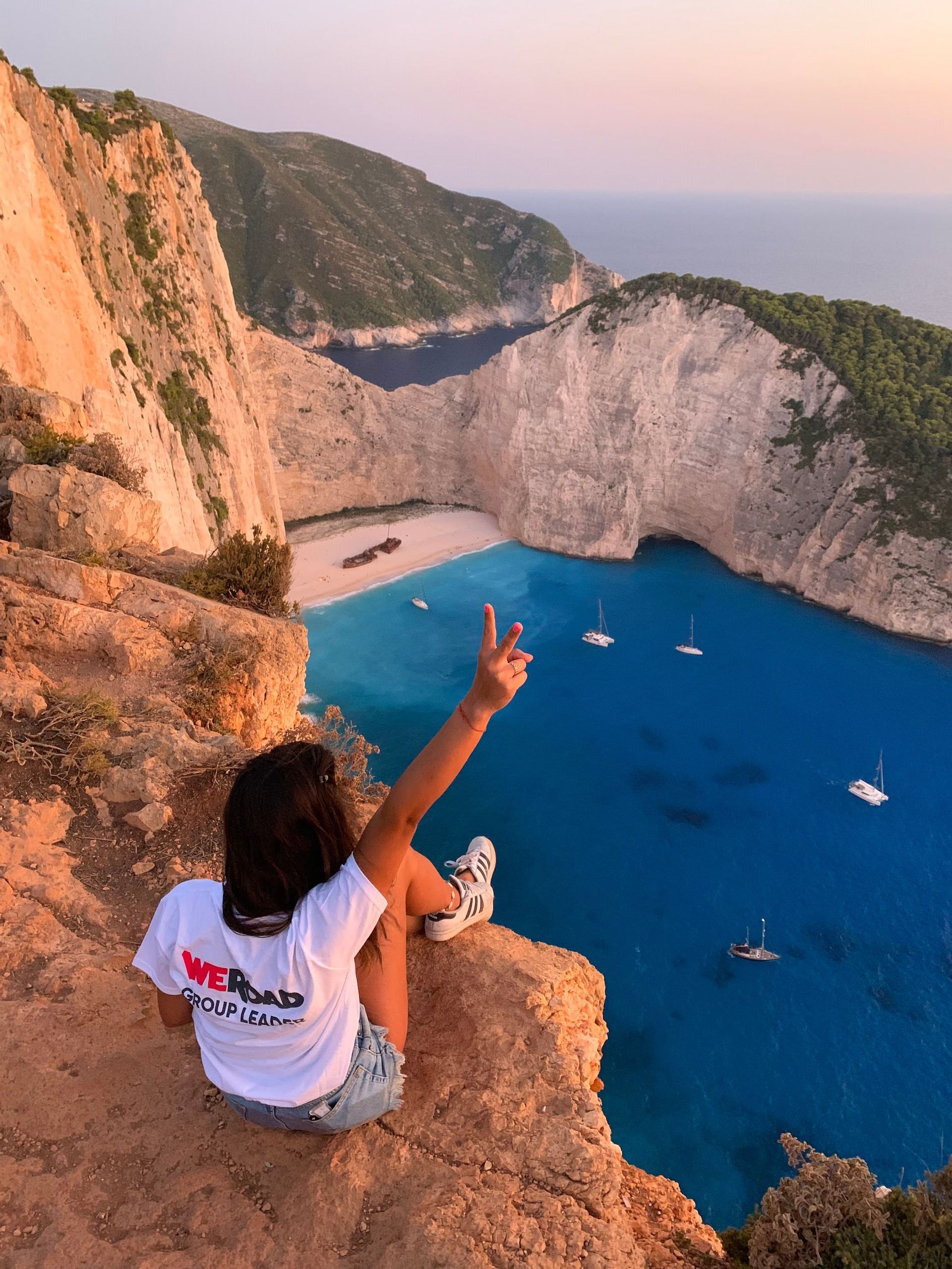 Un líder de grupo de WeRoad se sienta al borde de un acantilado con vistas a una cala con un naufragio en la playa y barcos en el agua turquesa.