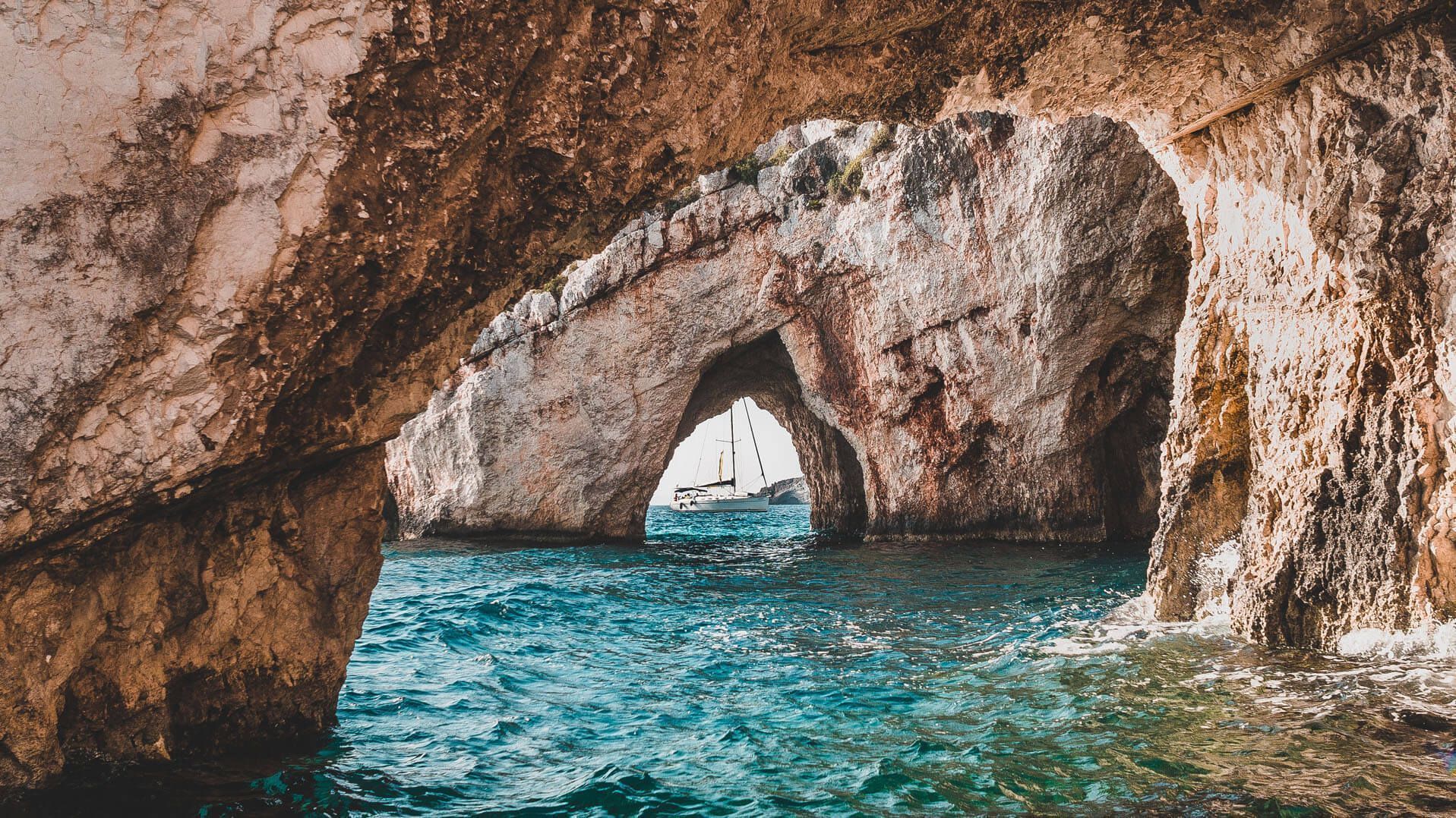 Velero blanco enmarcado por un arco de roca natural, visto desde el interior de una cueva marina sobre agua turquesa.