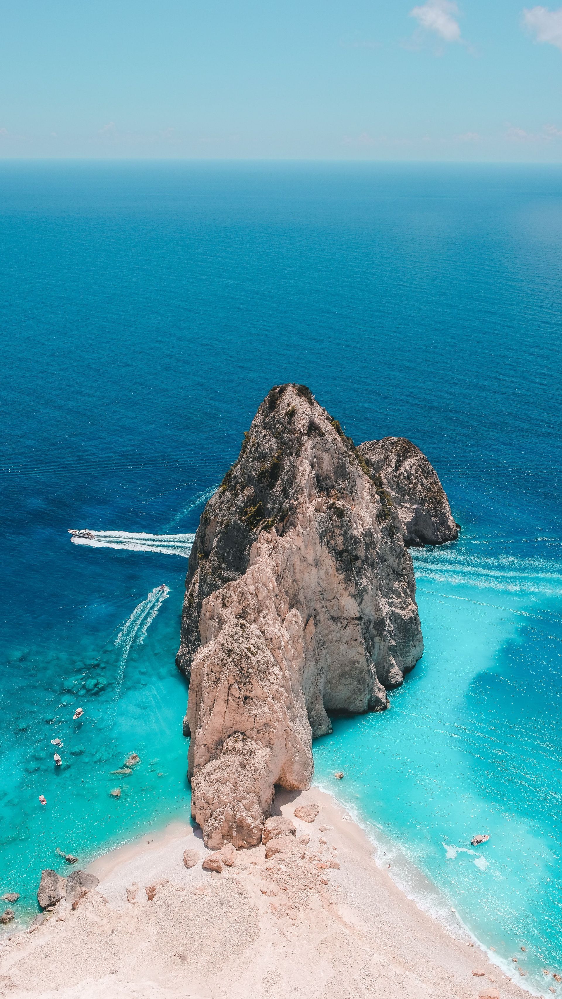 Una vista aerea di una grande formazione rocciosa su una spiaggia circondata da acqua turchese. Diverse barche navigano nel mare blu profondo nelle vicinanze.