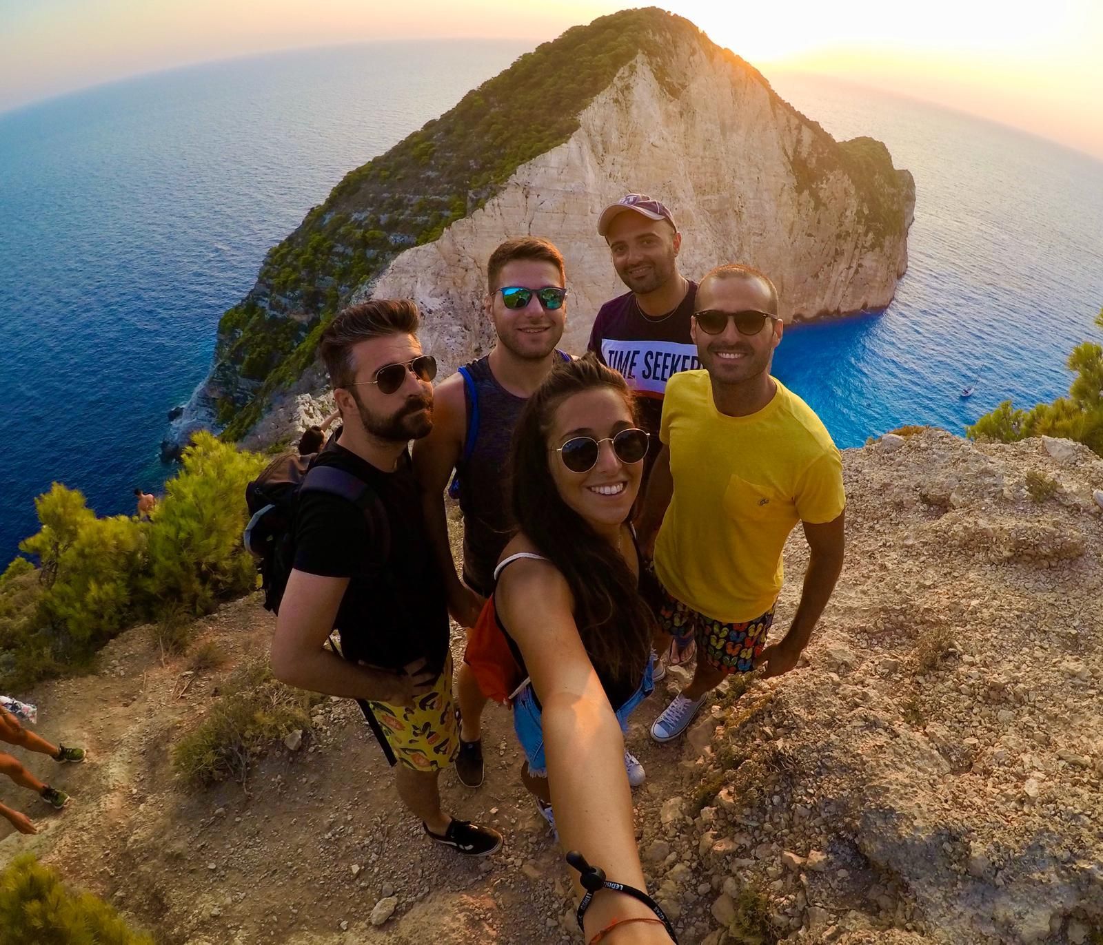 Un gruppo WeRoad si fa un selfie su una scogliera a picco su una costa rocciosa e il mare blu al tramonto.
