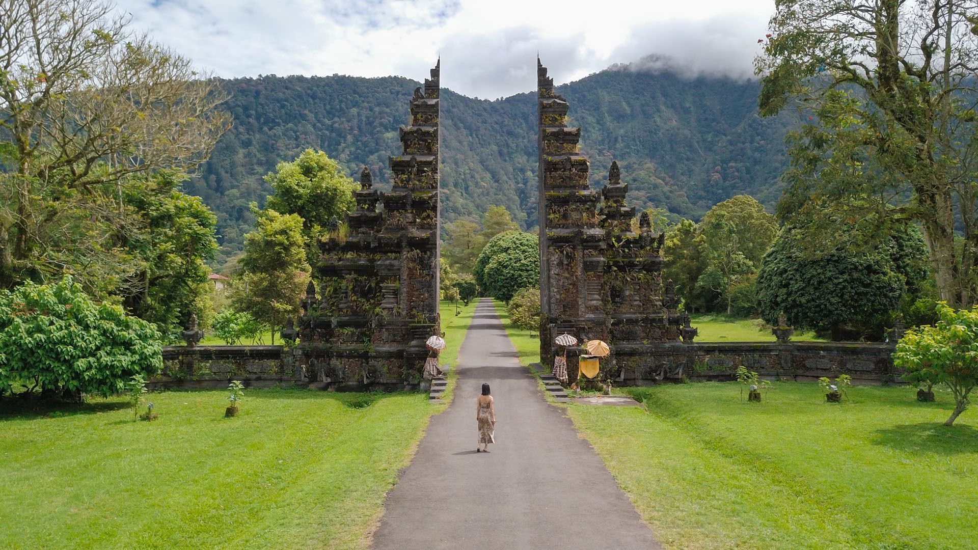 A woman seen from behind walks on a path towards an ornate split stone gate, with lush green trees and a mountain in the background.
