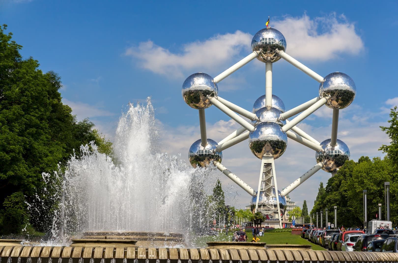 Le monument de l'Atomium se dresse derrière une grande fontaine d'eau dans un parc sous un ciel bleu avec des nuages épars.