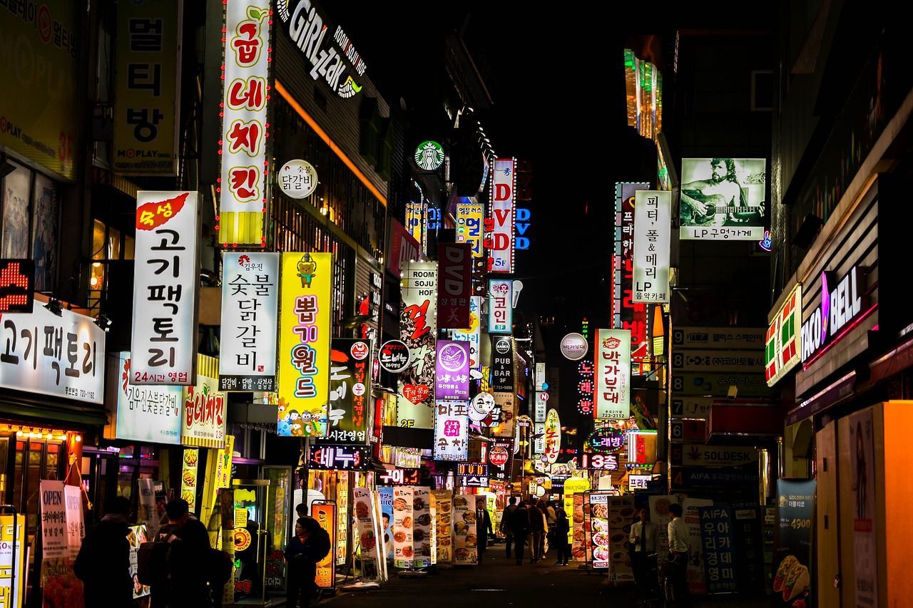 Una strada cittadina affollata di notte, illuminata da molte insegne al neon verticali e colorate, con scritte in coreano.