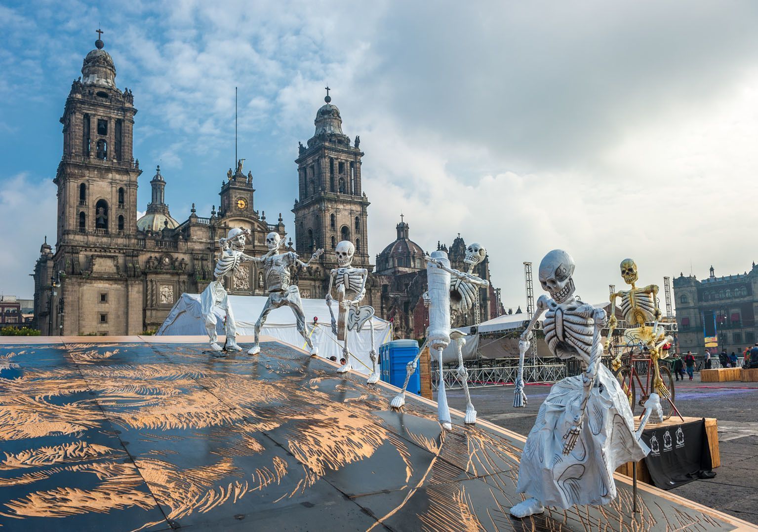 Varias figuras de esqueletos grandes para un festival se encuentran en una plaza frente a una gran catedral de dos torres bajo un cielo azul.