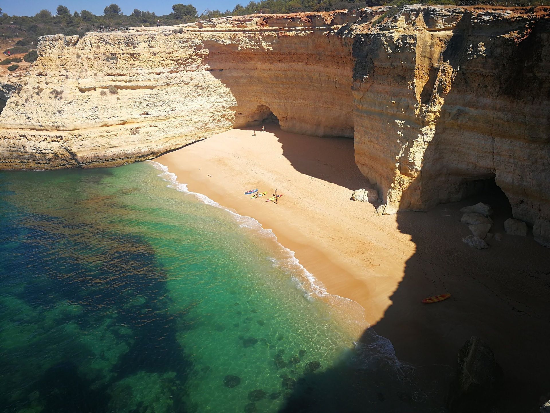 Ein abgelegener Sandstrand mit klarem, türkisfarbenem Wasser, umgeben von hohen Klippen mit Höhlen, aus der Vogelperspektive gesehen.