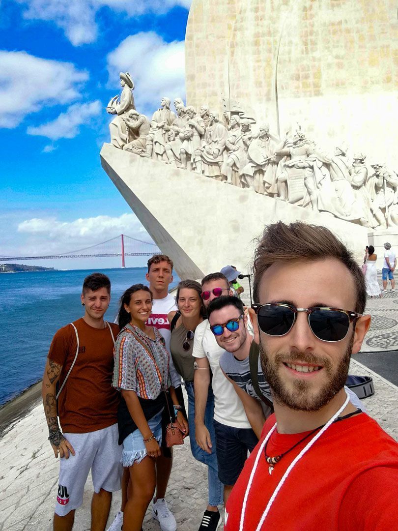 Eine WeRoad-Gruppe macht ein Selfie vor einem großen Steinmonument mit Brücke und Wasser im Hintergrund.