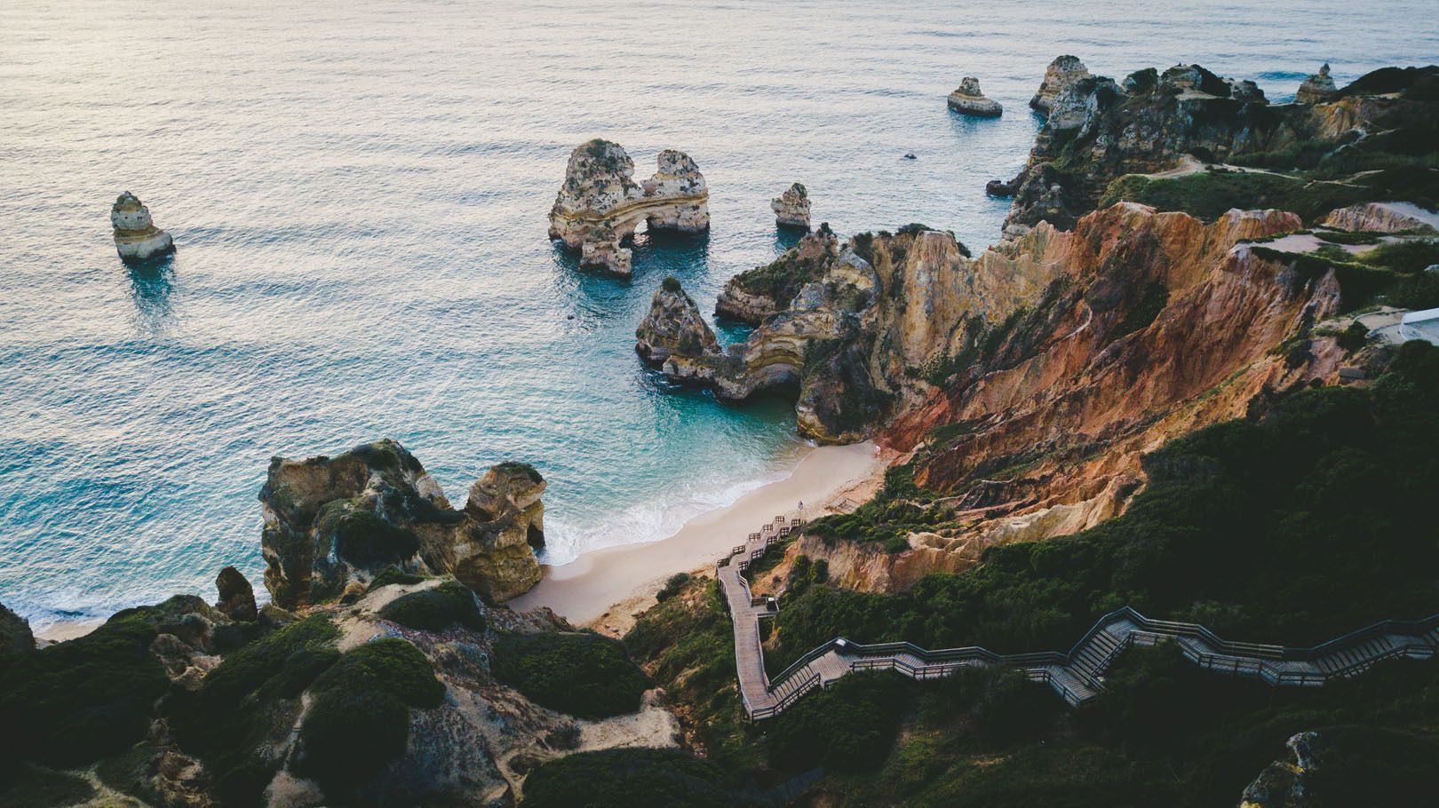 Eine Luftaufnahme einer Holztreppe, die sich einen grünen, felsigen Hang hinunter zu einem abgelegenen Sandstrand mit großen Felsnadeln im Wasser windet.