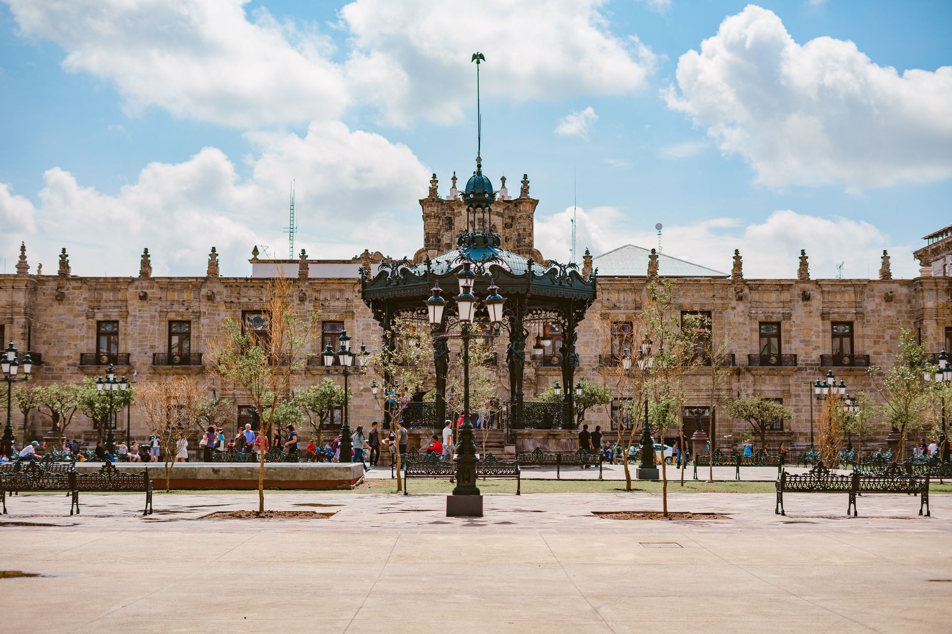 Un quiosco ornamentado y una farola antigua en una gran plaza pública frente a un edificio histórico de piedra bajo un cielo nublado.