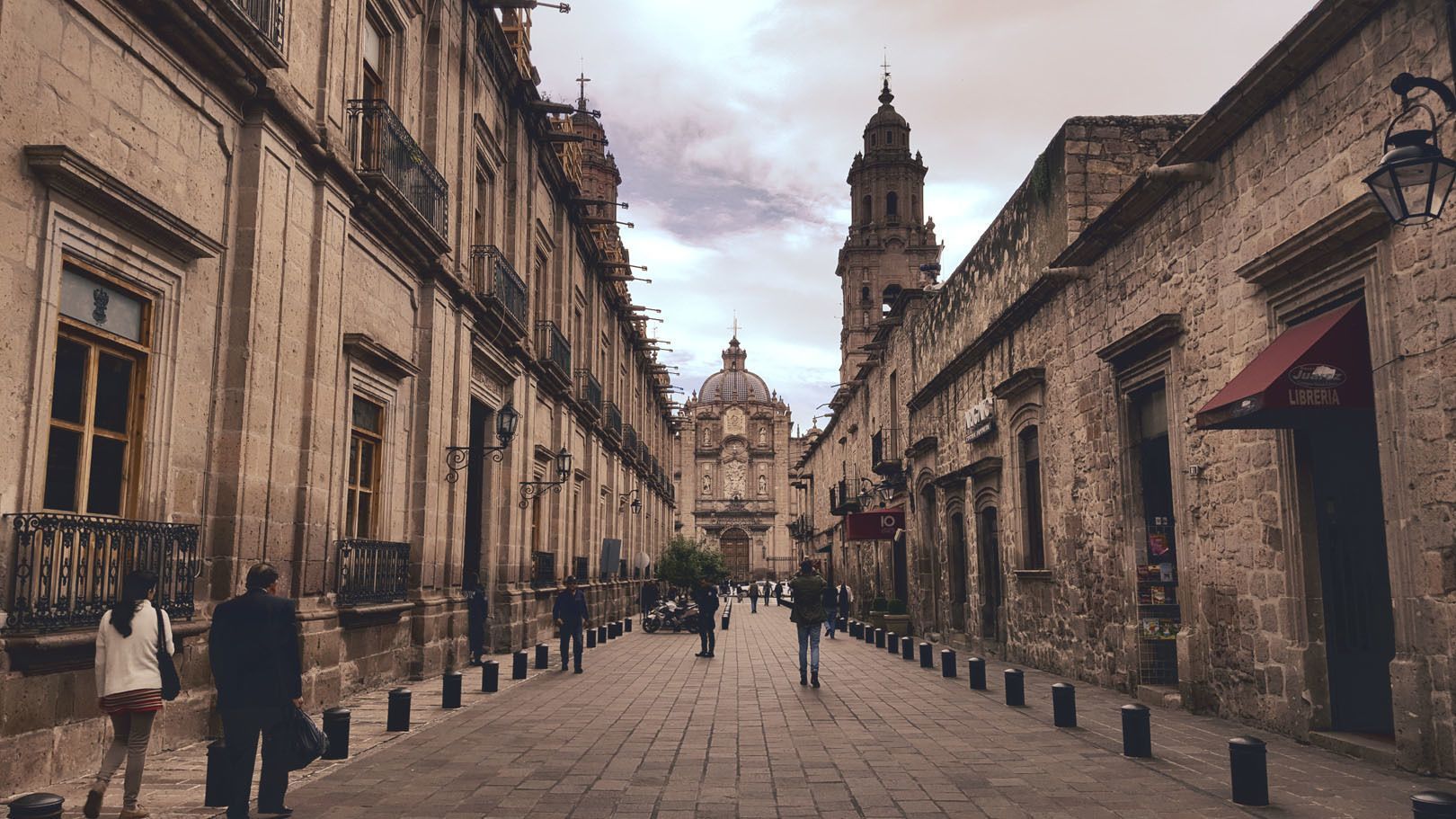Una vista de una calle peatonal adoquinada bordeada de edificios históricos de piedra, con una gran catedral y un campanario al final.