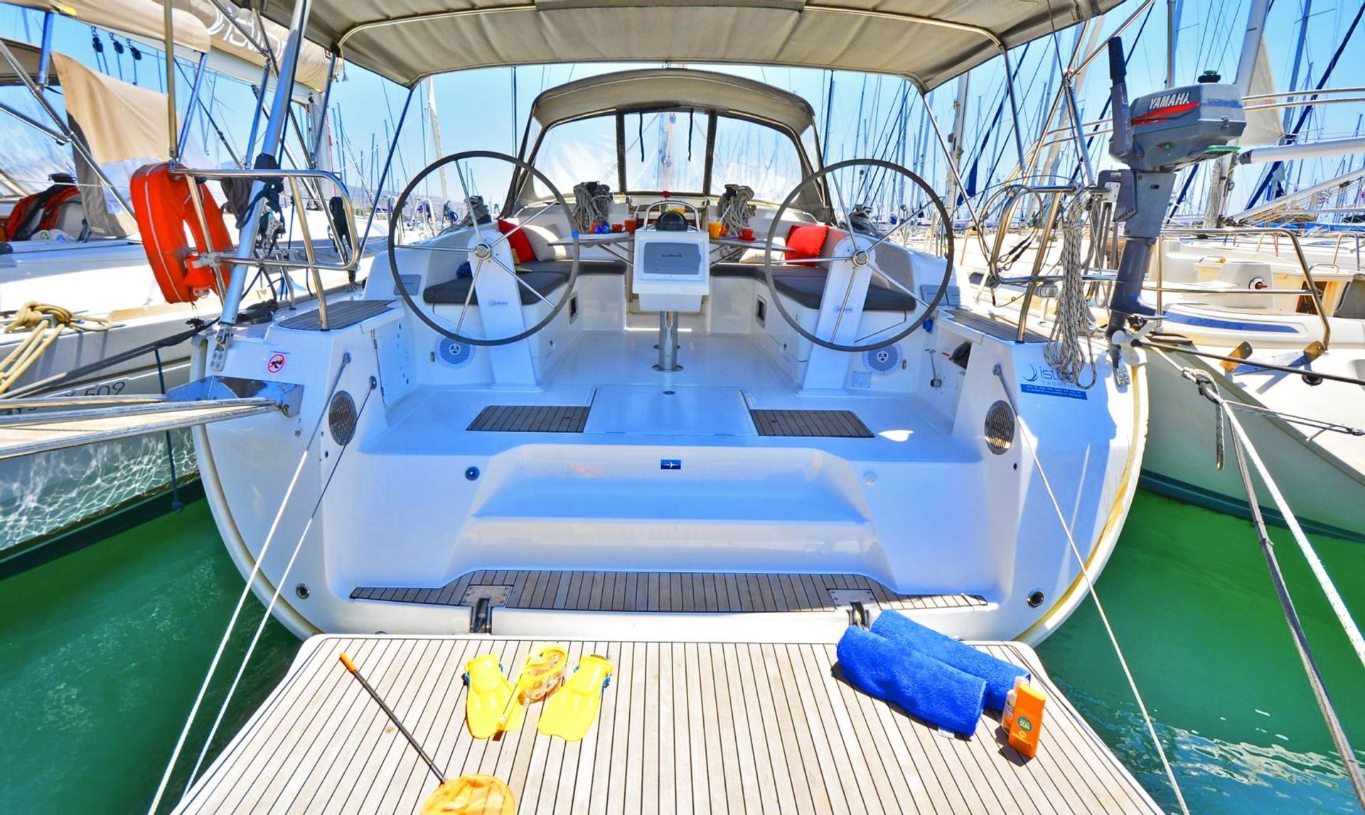 La poppa e il pozzetto di una barca a vela con doppie ruote di governo, ormeggiata in una marina con pinne e asciugamani sul ponte.