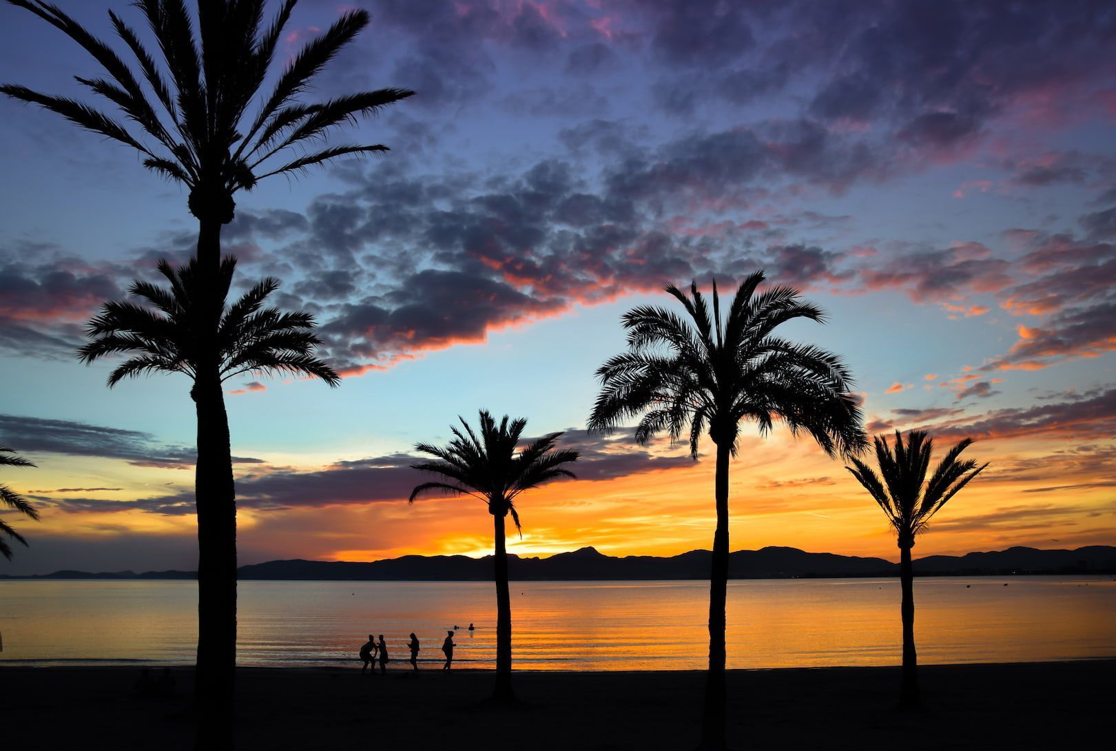 Palme e persone in silhouette su una spiaggia, contro un tramonto colorato sull'acqua, con montagne all'orizzonte.