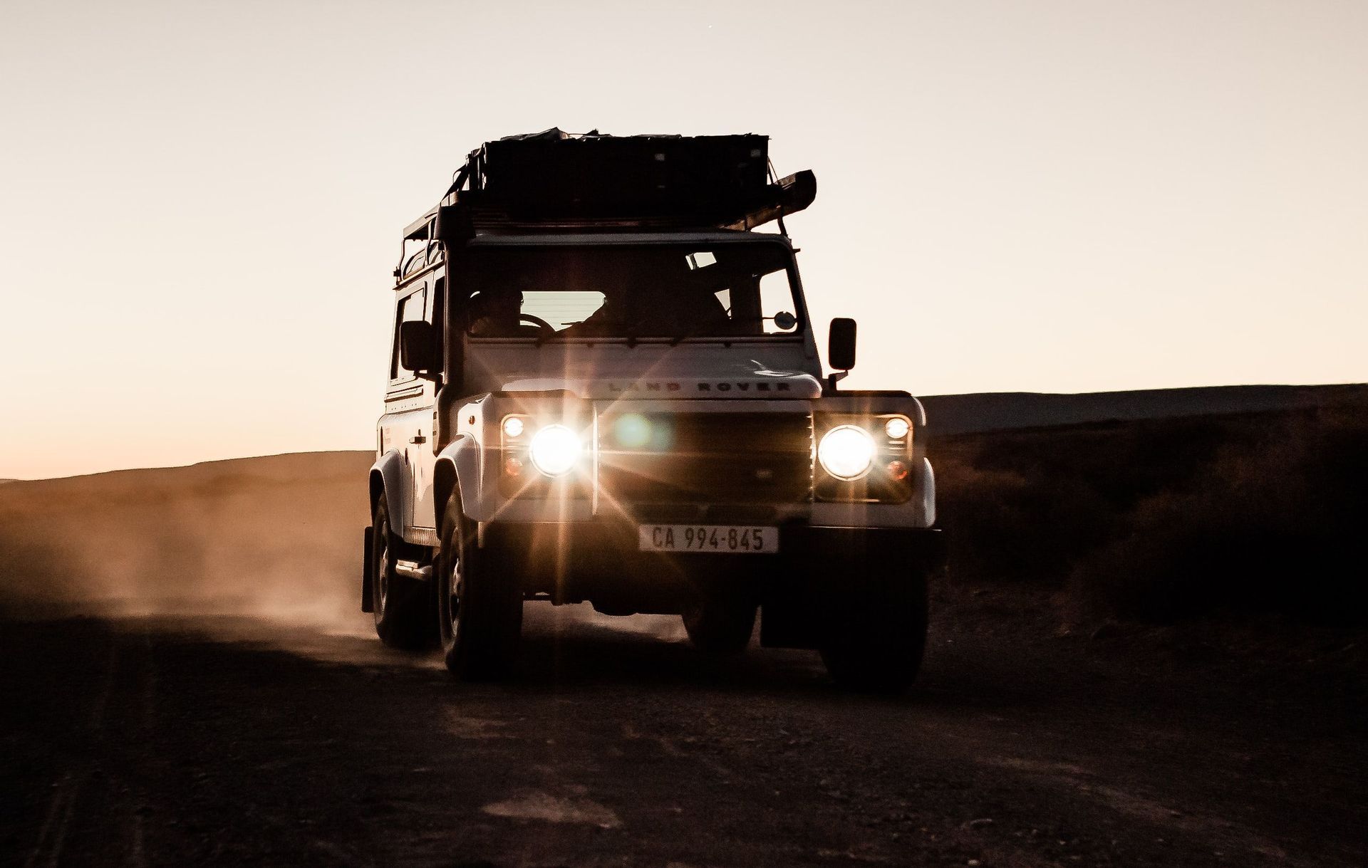 Ein weißer Land Rover mit einer WeRoad-Gruppenreise fährt bei Sonnenuntergang mit eingeschalteten Scheinwerfern auf einer staubigen Straße.