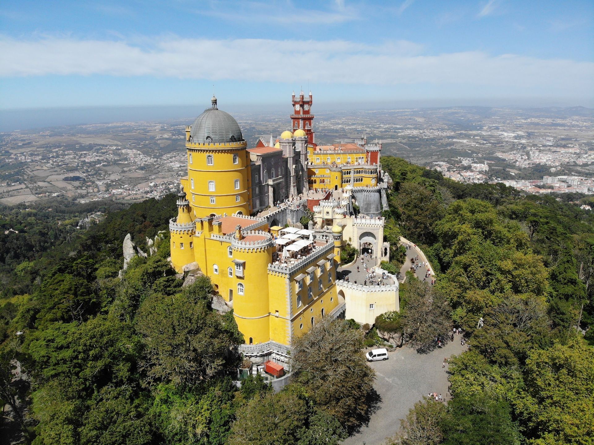 Un château coloré jaune et rouge avec des tours se dresse sur une colline boisée surplombant une ville tentaculaire sous un ciel bleu.