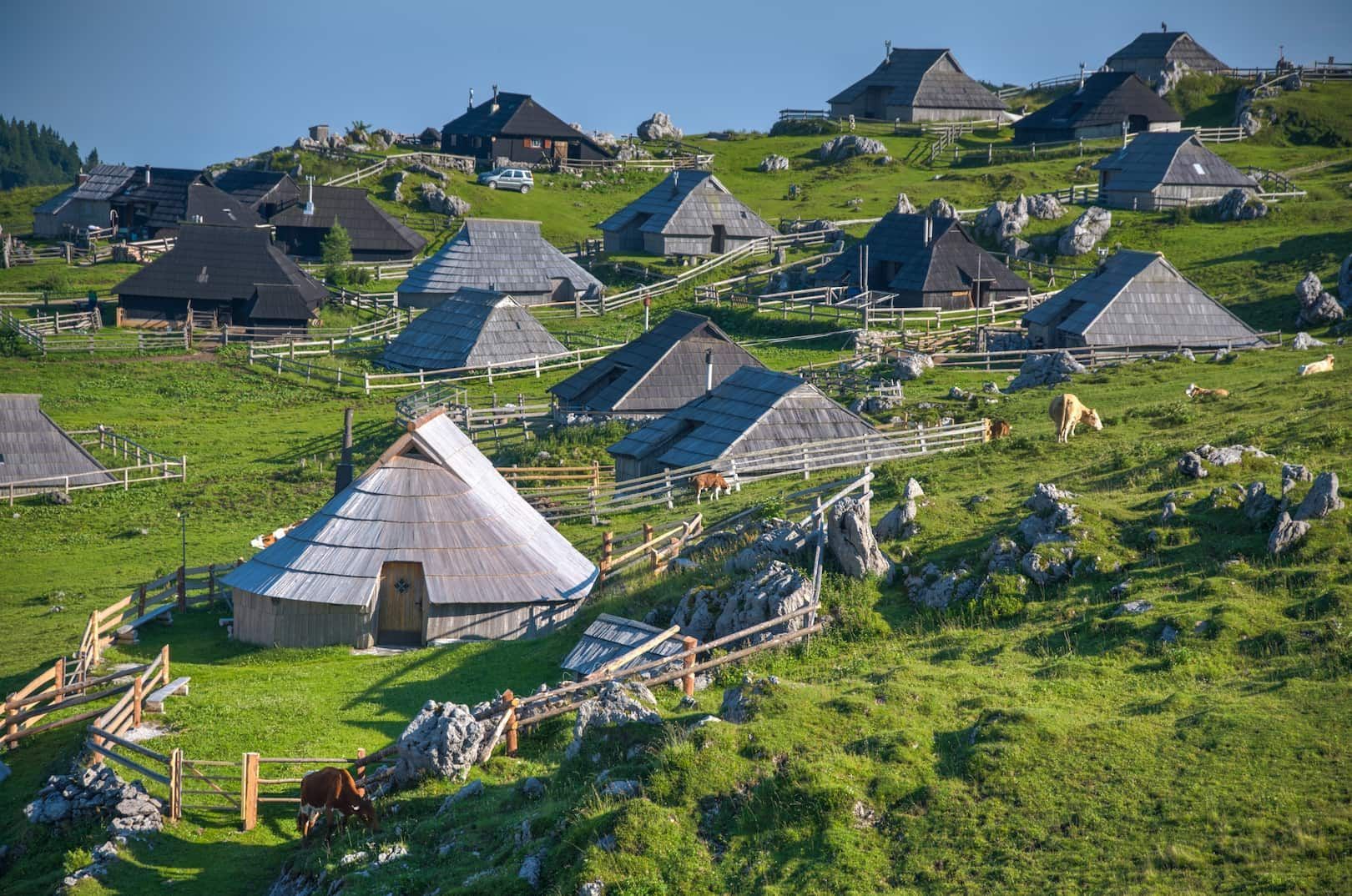 Un pueblo de cabañas de madera tradicionales con techos de tejas se dispersa por una ladera de montaña verde y soleada con vacas pastando.