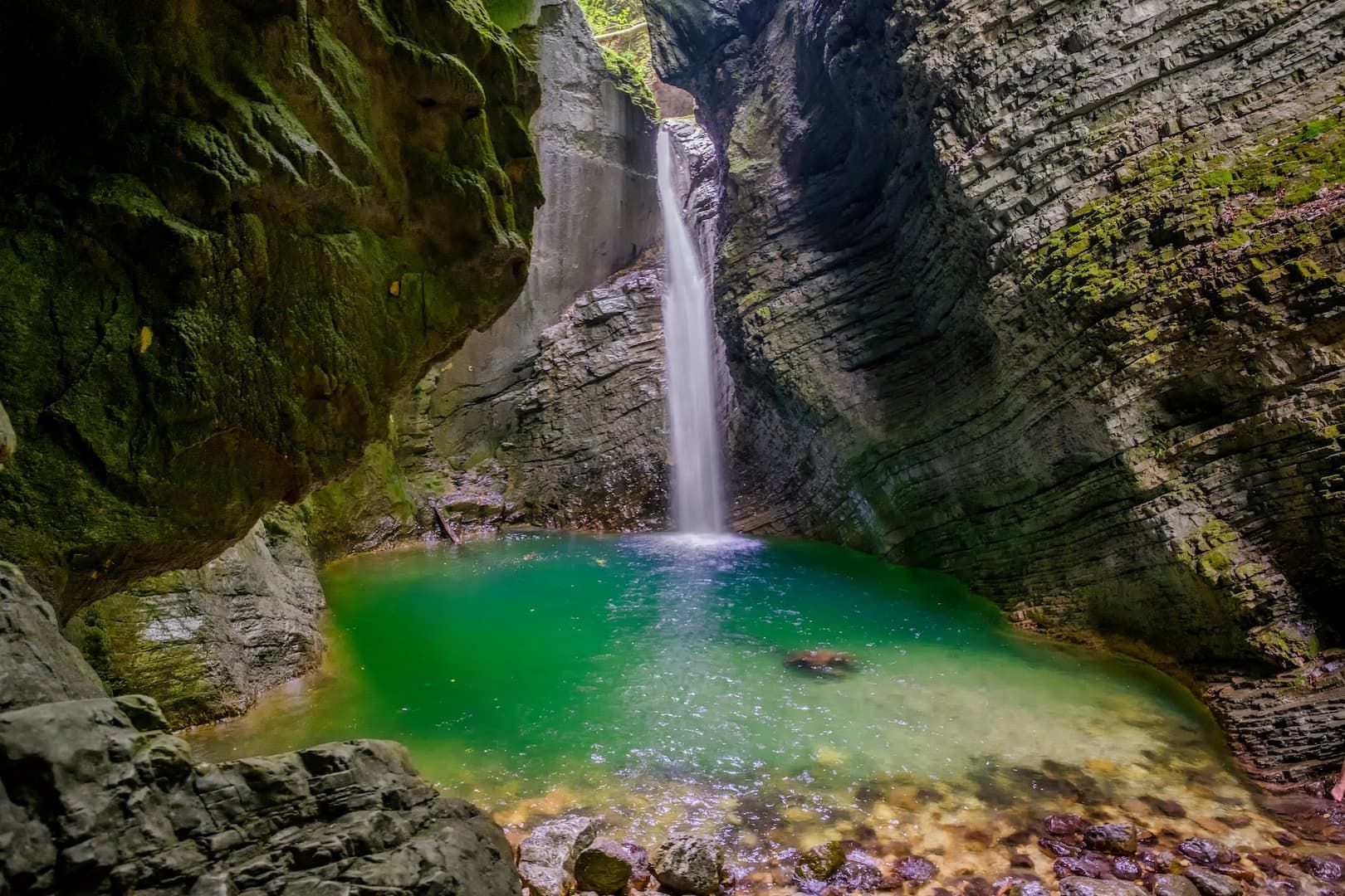 Ein Wasserfall stürzt in ein leuchtend türkises Wasserbecken, umgeben von steilen, moosbewachsenen Felswänden in einer Schlucht.