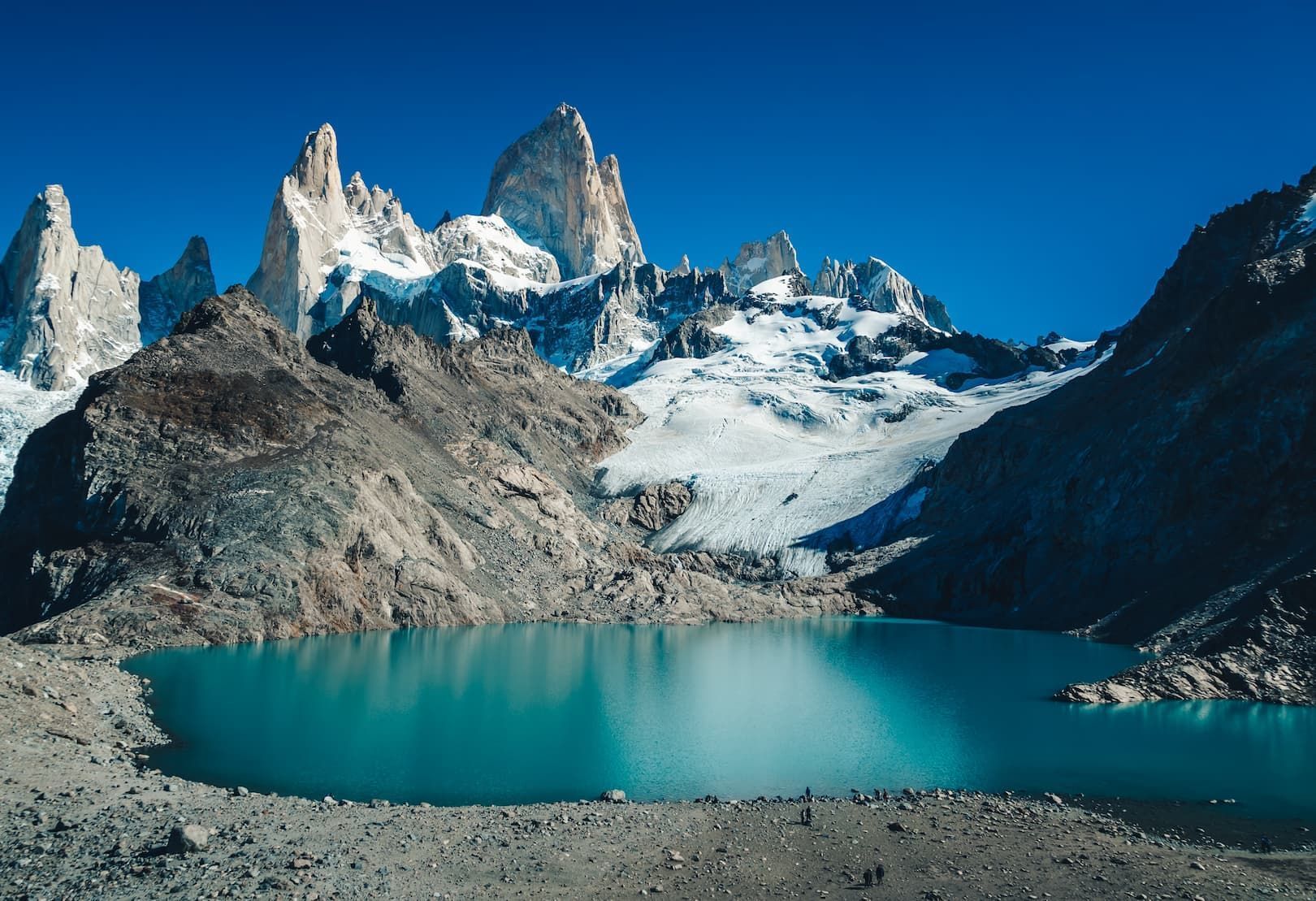 Un grupo de WeRoad camina por la orilla rocosa de un lago turquesa, al pie de montañas escarpadas y nevadas, bajo un cielo azul claro.