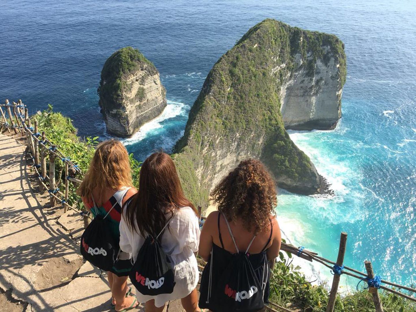 Tres mujeres de un viaje en grupo de WeRoad están de pie en un sendero junto a un acantilado, observando grandes formaciones rocosas en un océano turquesa.