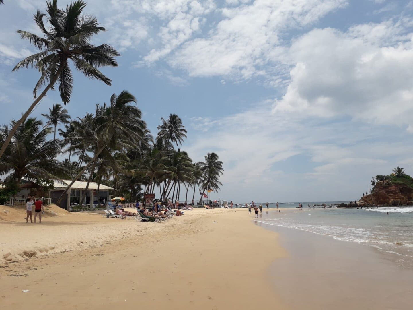 Une journée ensoleillée sur une plage de sable fin, où des gens se détendent et se promènent au bord de l'océan, bordé de grands palmiers et d'un petit bâtiment.