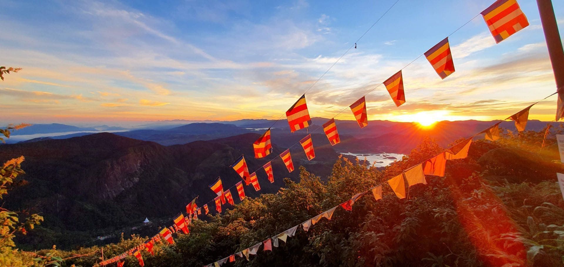 Strings of colorful Buddhist prayer flags hang over a lush mountain valley as the sun rises on the horizon, illuminating the landscape.