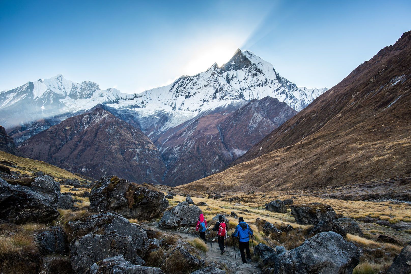Un groupe WeRoad de trois personnes en randonnée avec des bâtons sur un sentier rocheux, vers une imposante montagne enneigée au lever du soleil.